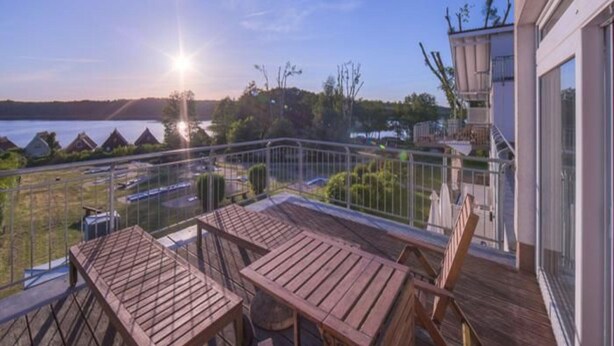 A wooden deck with tables and chairs overlooking a body of water and a forested area. - Ferienwohnung im Aparthotel Seepanorama (Mirow)