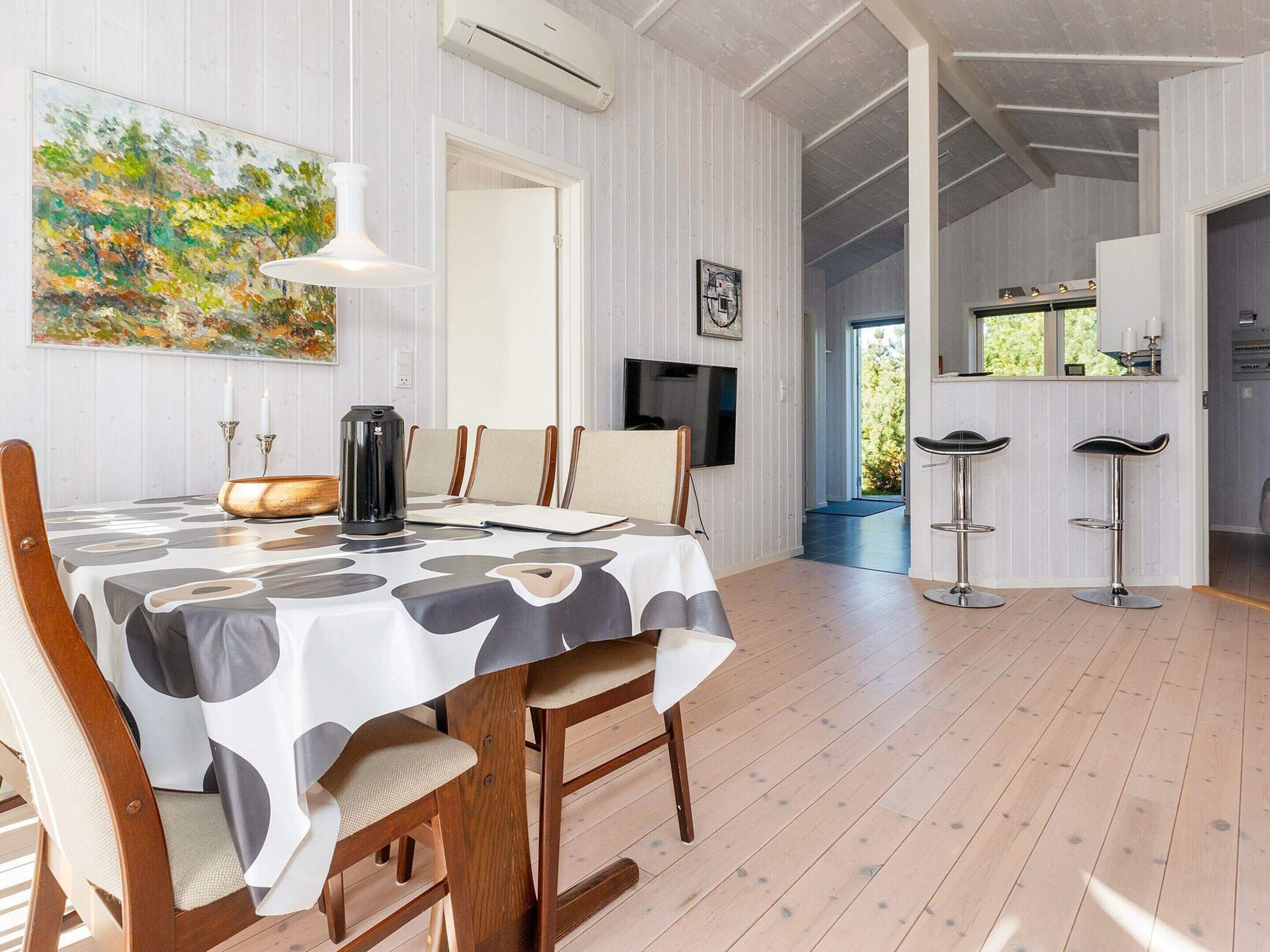 Un coin repas avec une table dressée pour quatre personnes, des chaises en bois, un luminaire suspendu moderne, un tableau au mur et un téléviseur.