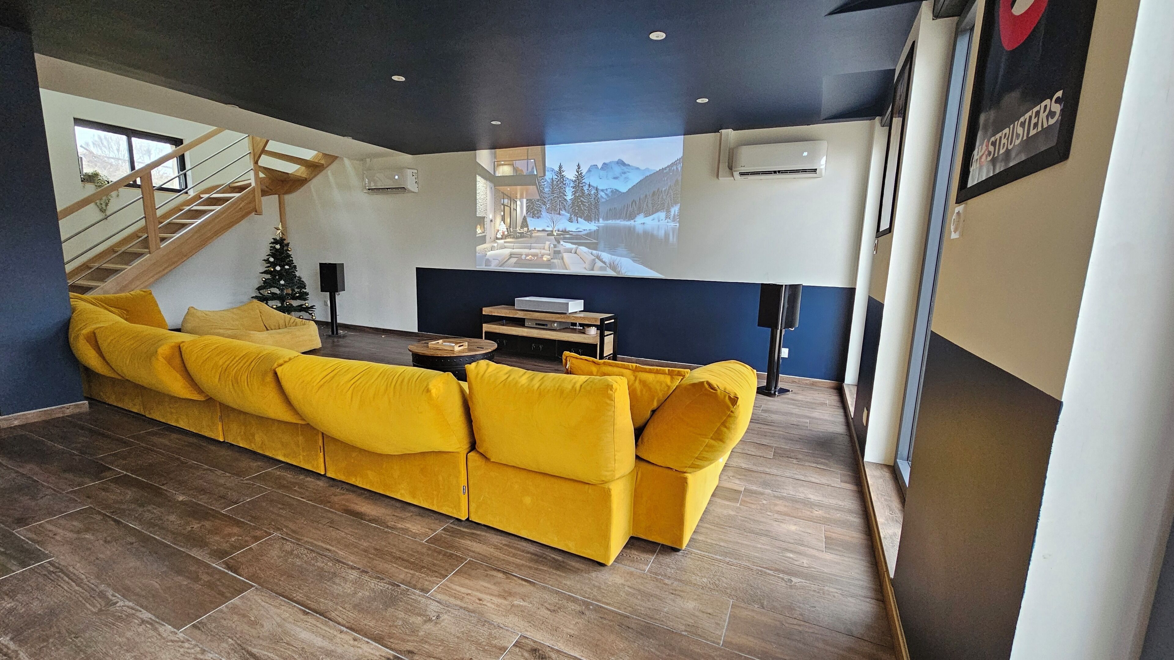 Una sala de estar moderna con un sofá seccional amarillo, una escalera y una gran pantalla de televisión que muestra un paisaje nevado.