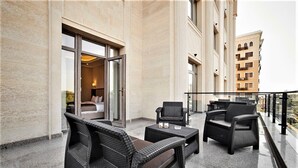 Ein Balkon mit Korbmöbeln, eine Glastür, die zu einem Zimmer führt, und ein Blick auf ein Gebäude im Hintergrund.