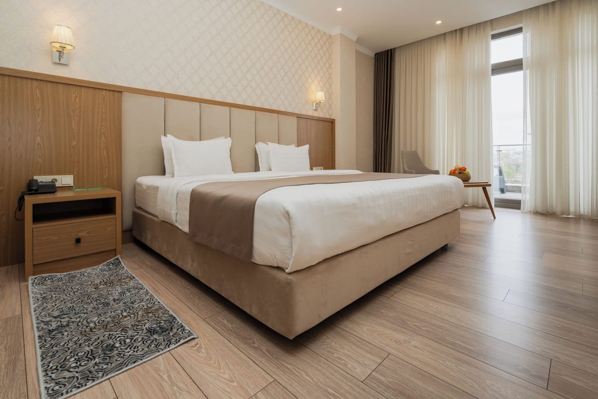 A modern hotel room with a large bed, wooden headboard, bedside tables, and a view of the outdoors through a large window.