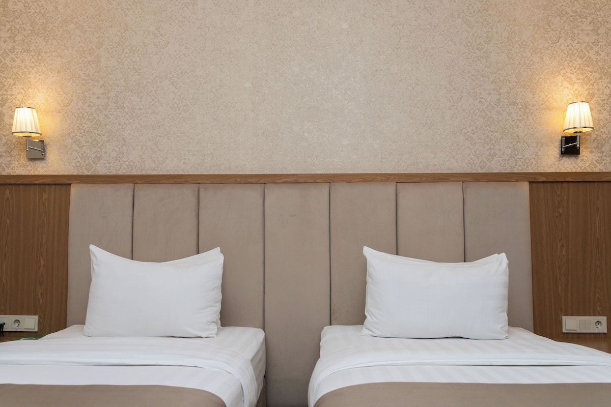 A hotel room with two beds, wooden headboard, wall-mounted lamps, and a textured wall.