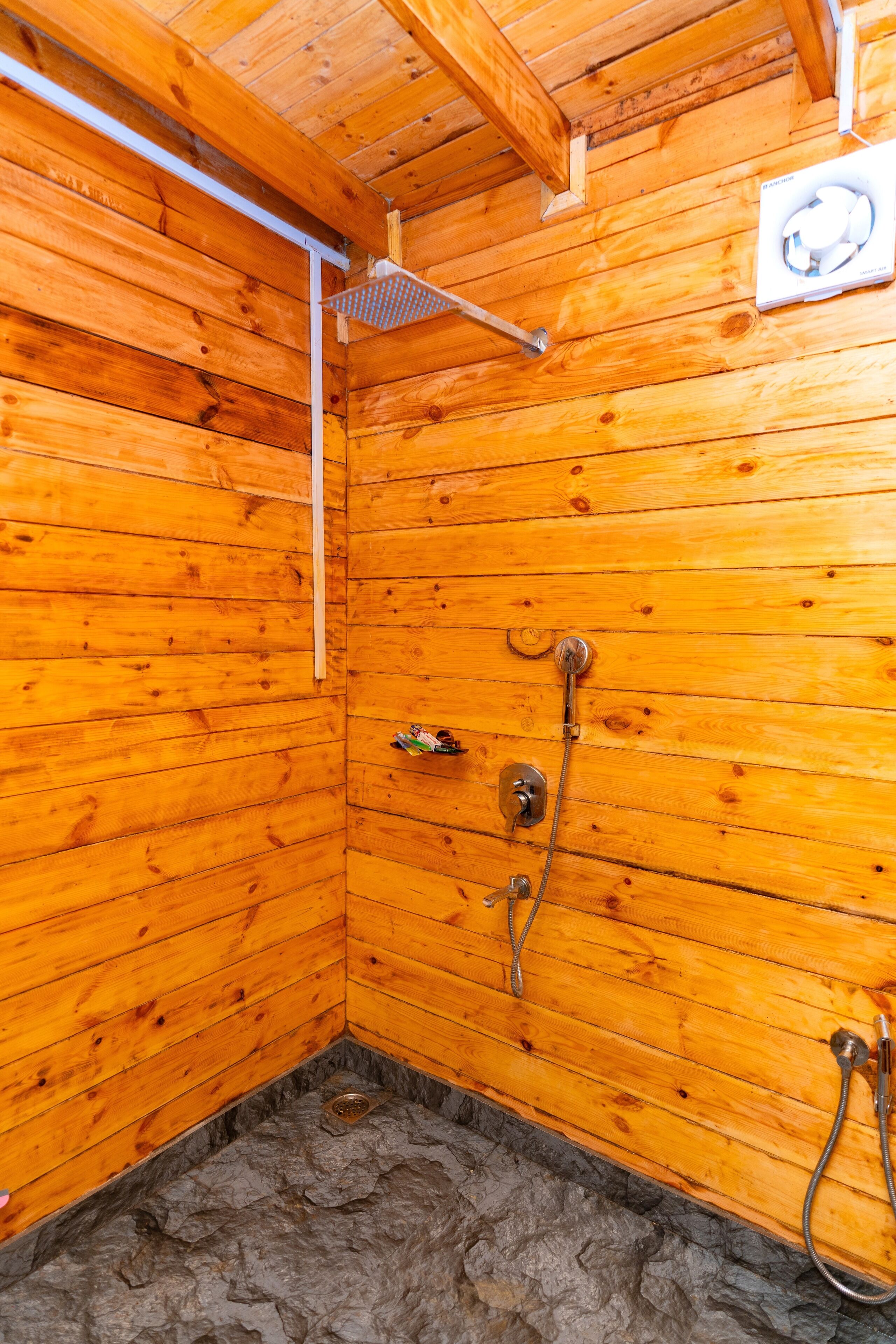 A wooden cabin with a shower area, including a showerhead, a handheld shower, and a wall-mounted fan.