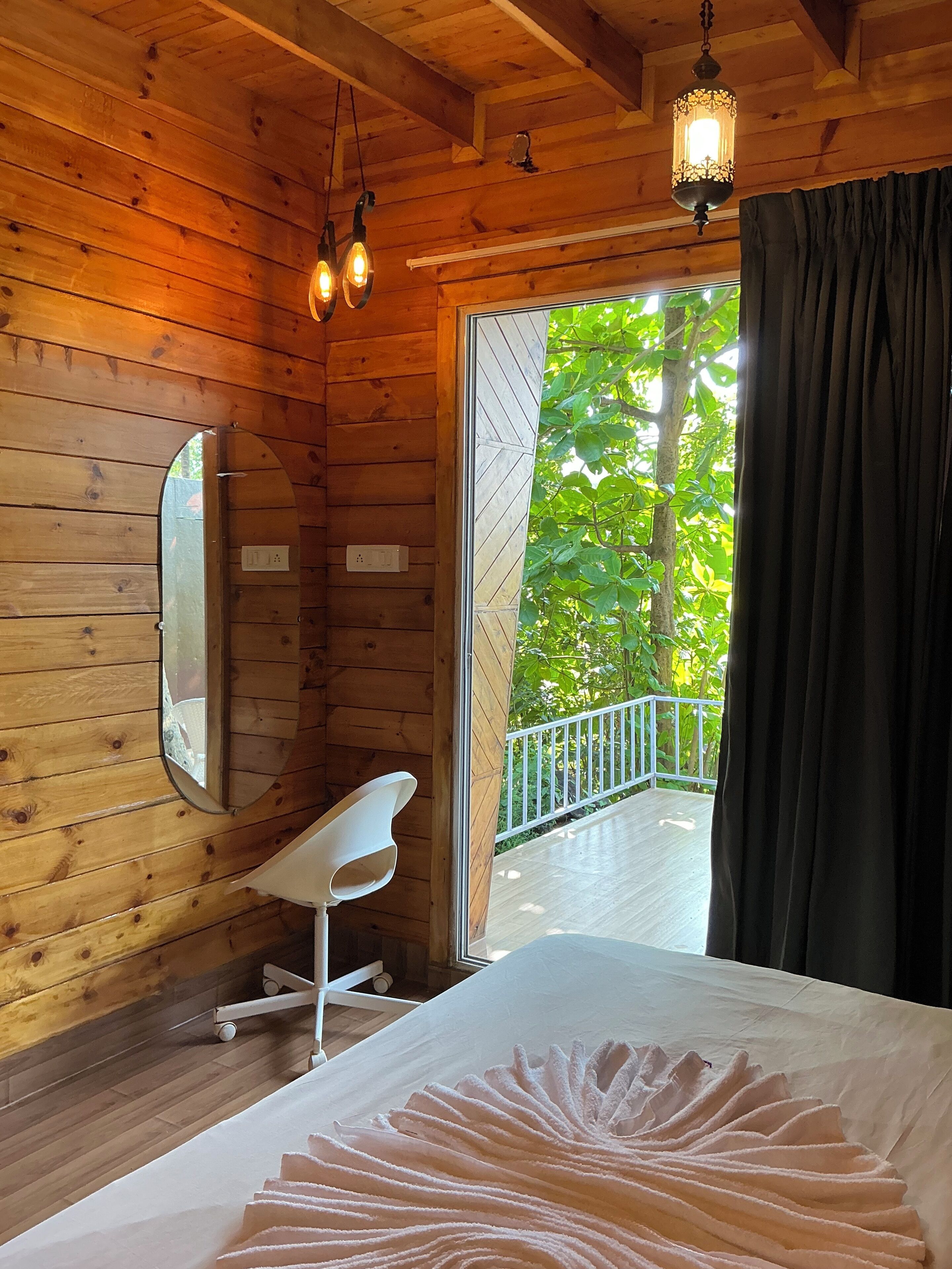 A wooden cabin room with a bed, a white chair, a mirror, and a balcony with a railing.