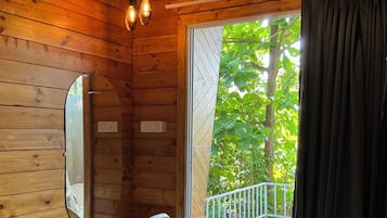 A wooden cabin room with a bed, a white chair, a mirror, and a balcony with a railing.