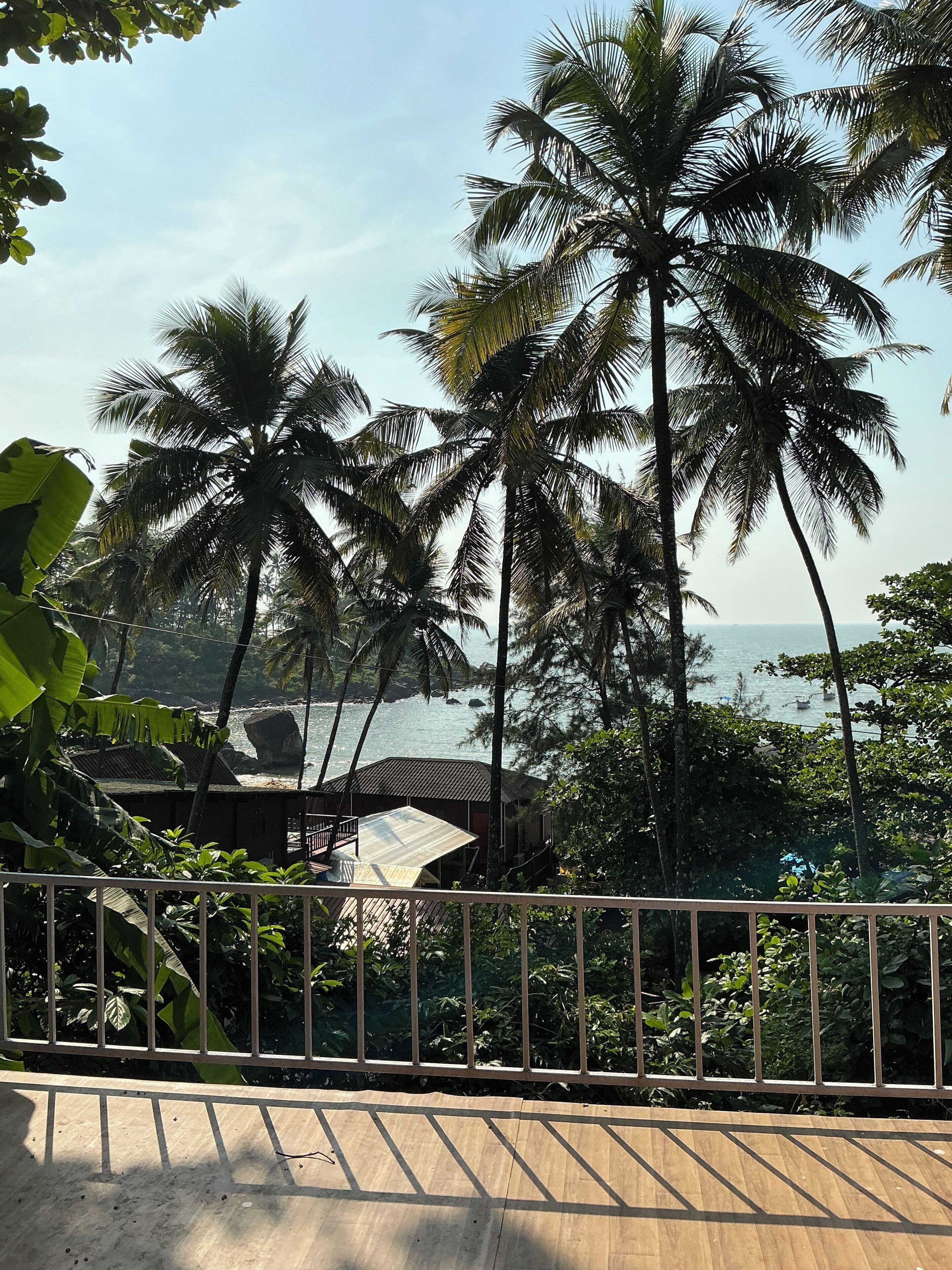 A wooden deck with a metal railing, overlooking a beach with palm trees and a building with a corrugated metal roof.