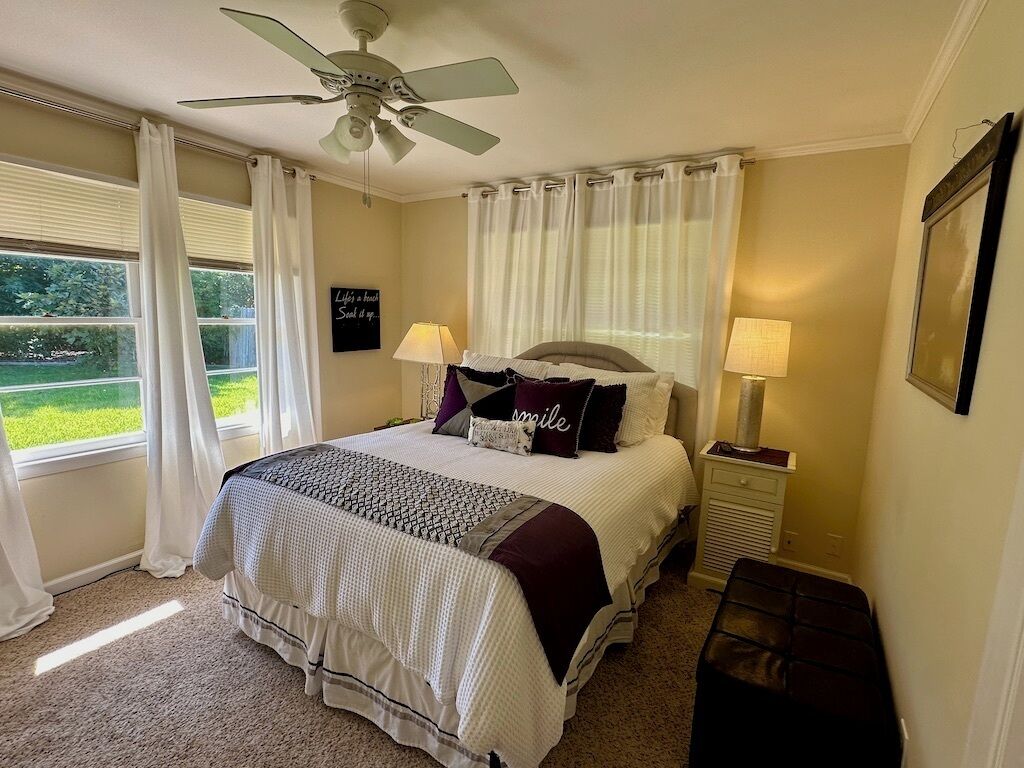 Une chambre à coucher avec un lit, deux lampes de chevet, un ventilateur de plafond, une fenêtre avec des rideaux et une vue sur la verdure à l’extérieur.