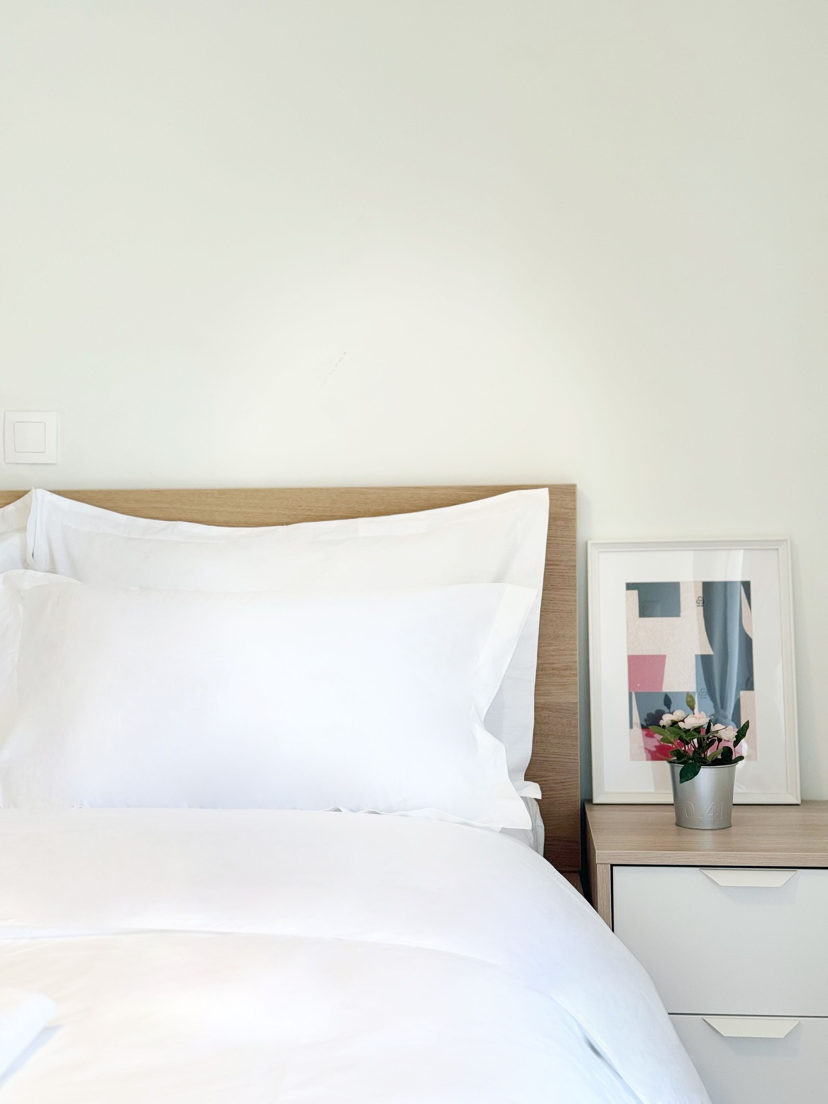 Una cama bien tendida con sábanas blancas, un cabecero de madera, una mesita de noche con un cuadro enmarcado y una pequeña planta en maceta.