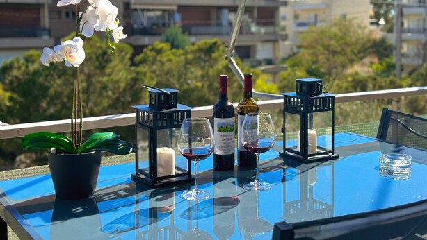 A balcony with a table set for a meal, featuring a potted plant, candles, wine bottles, glasses, and lanterns. - Blue Coast Residence Glyfada (Glyfada)