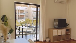 A modern living room with a dining area, a TV, and a balcony with a view of buildings. - Blue Coast Residence Glyfada (Glyfada)