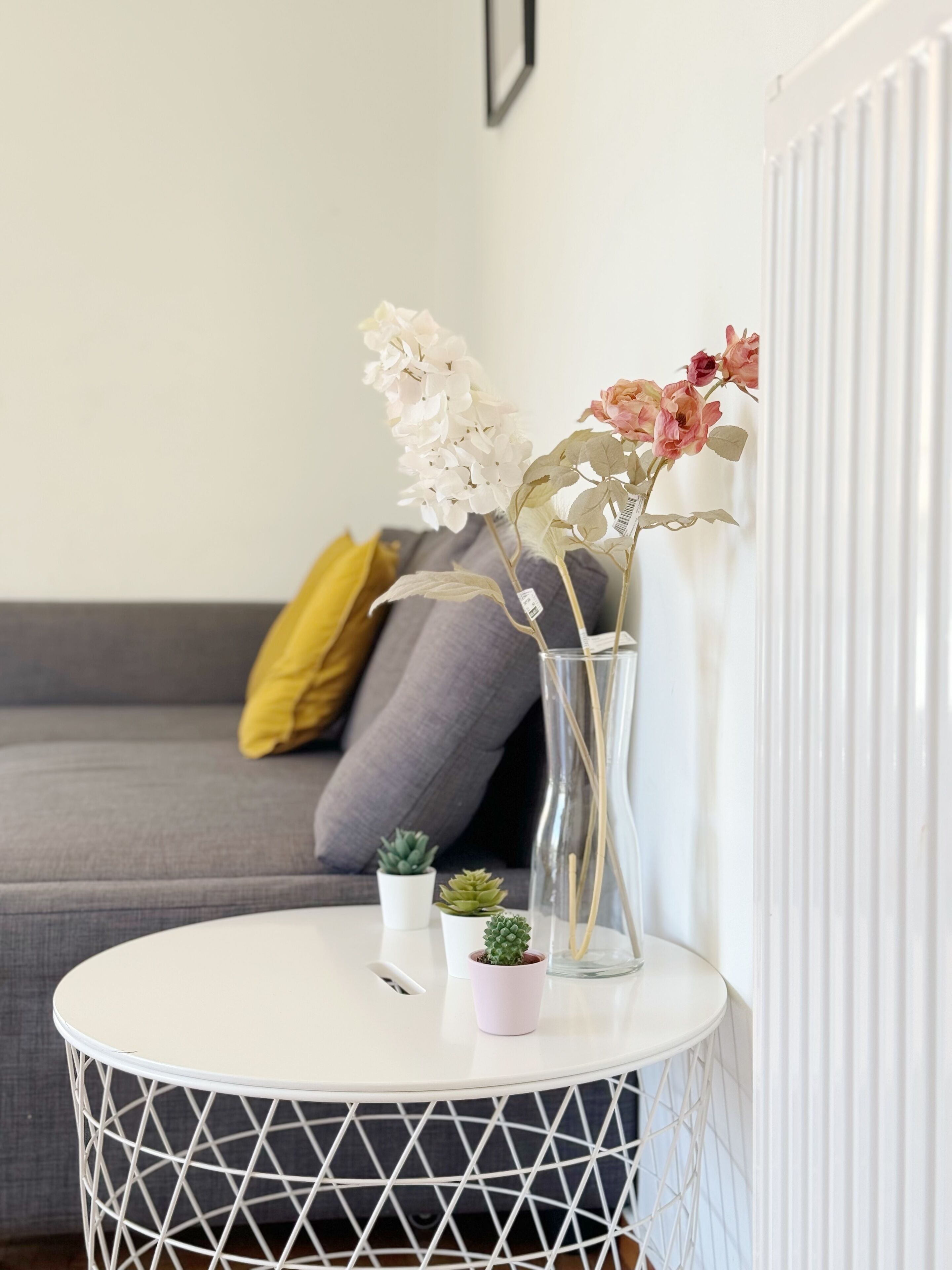 A round white table with a geometric pattern, a clear glass vase with dried flowers, and small potted plants on a grey sofa with cushions.