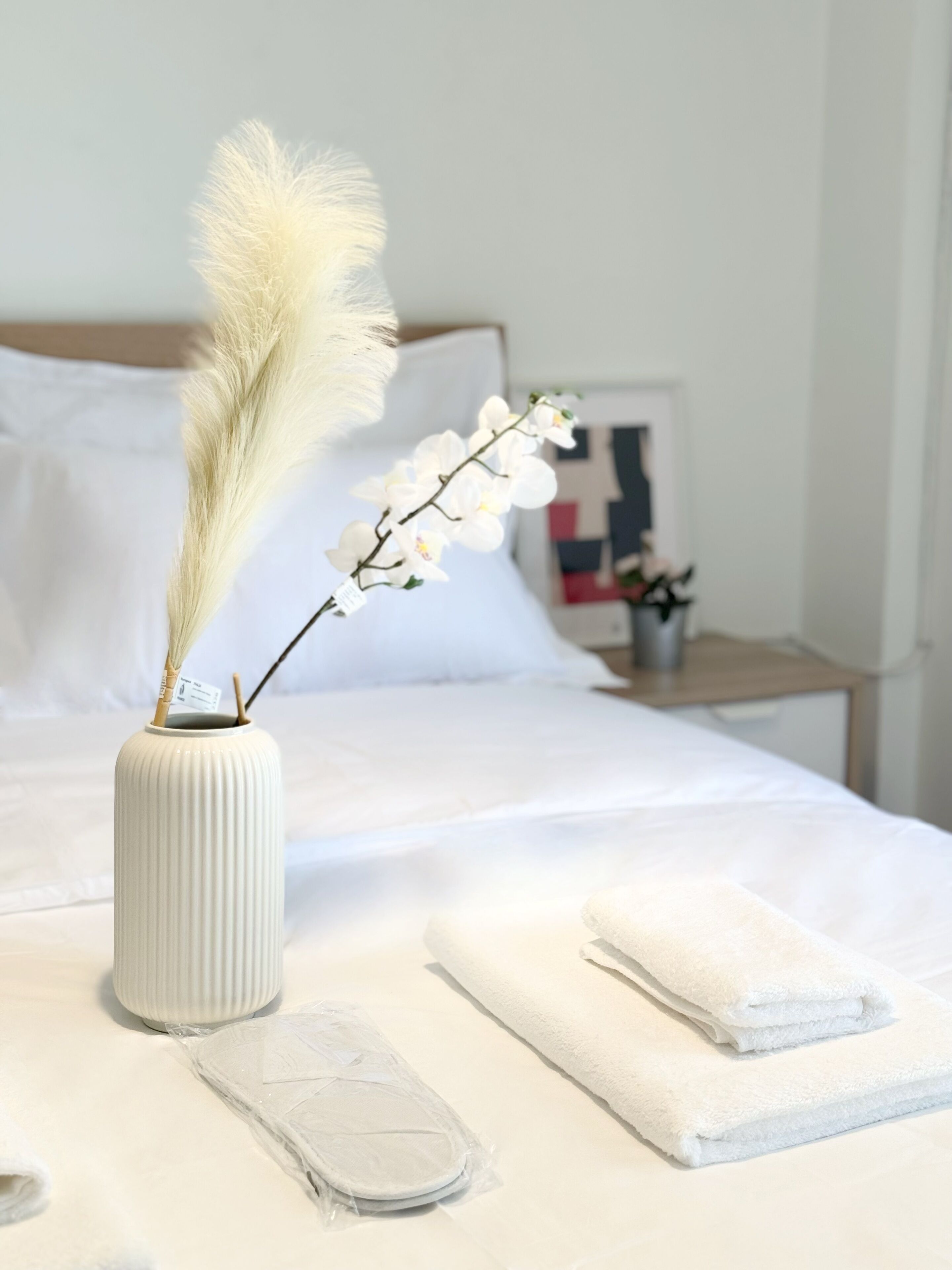 Una cama bien hecha con sábanas blancas, un jarrón blanco con una rama de flores blancas y un par de toallas blancas dobladas sobre la cama.