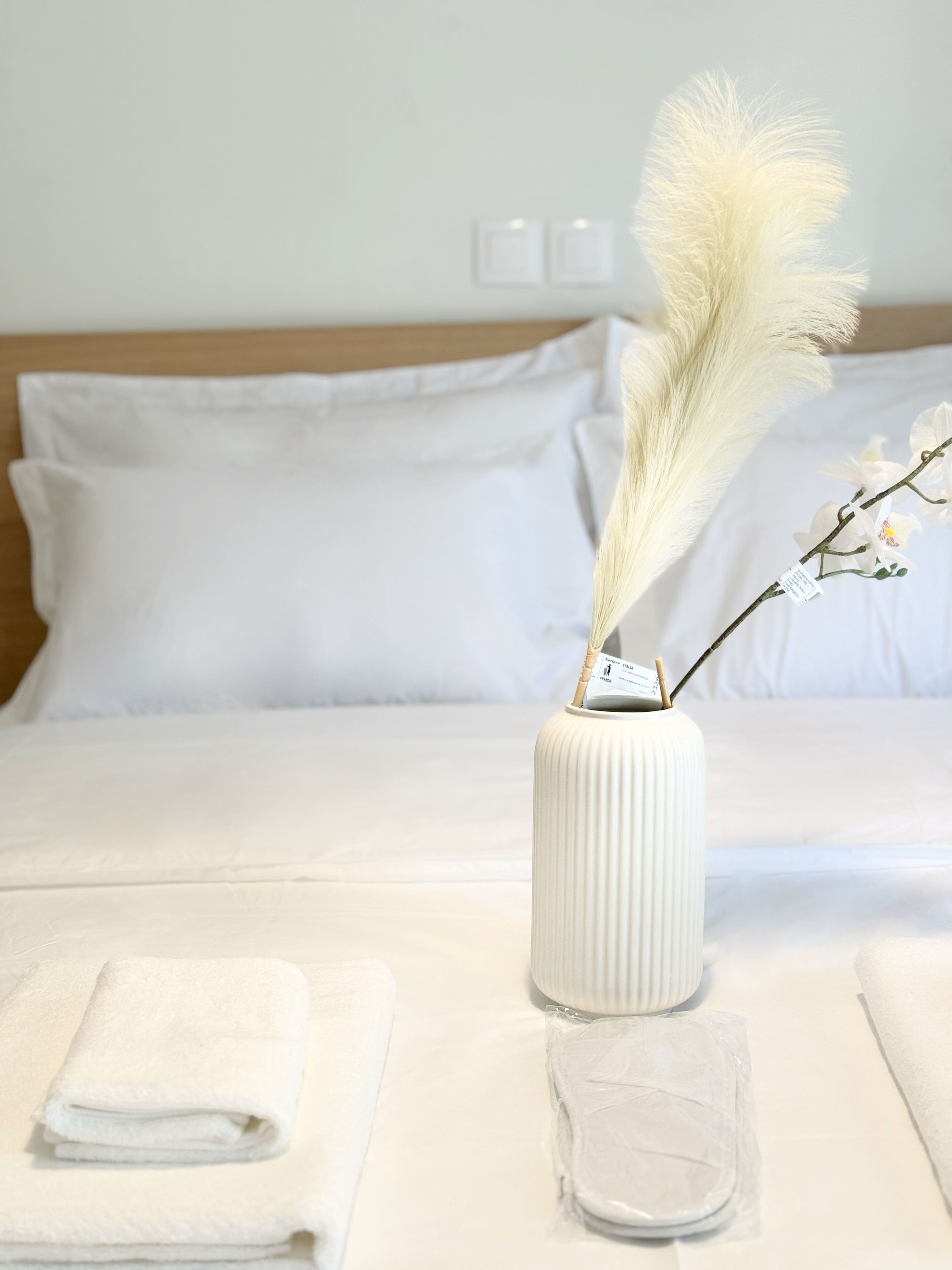 Una cama bien tendida con ropa blanca, un jarrón blanco con una rama y flores blancas, y una toalla blanca doblada sobre la mesita de noche.