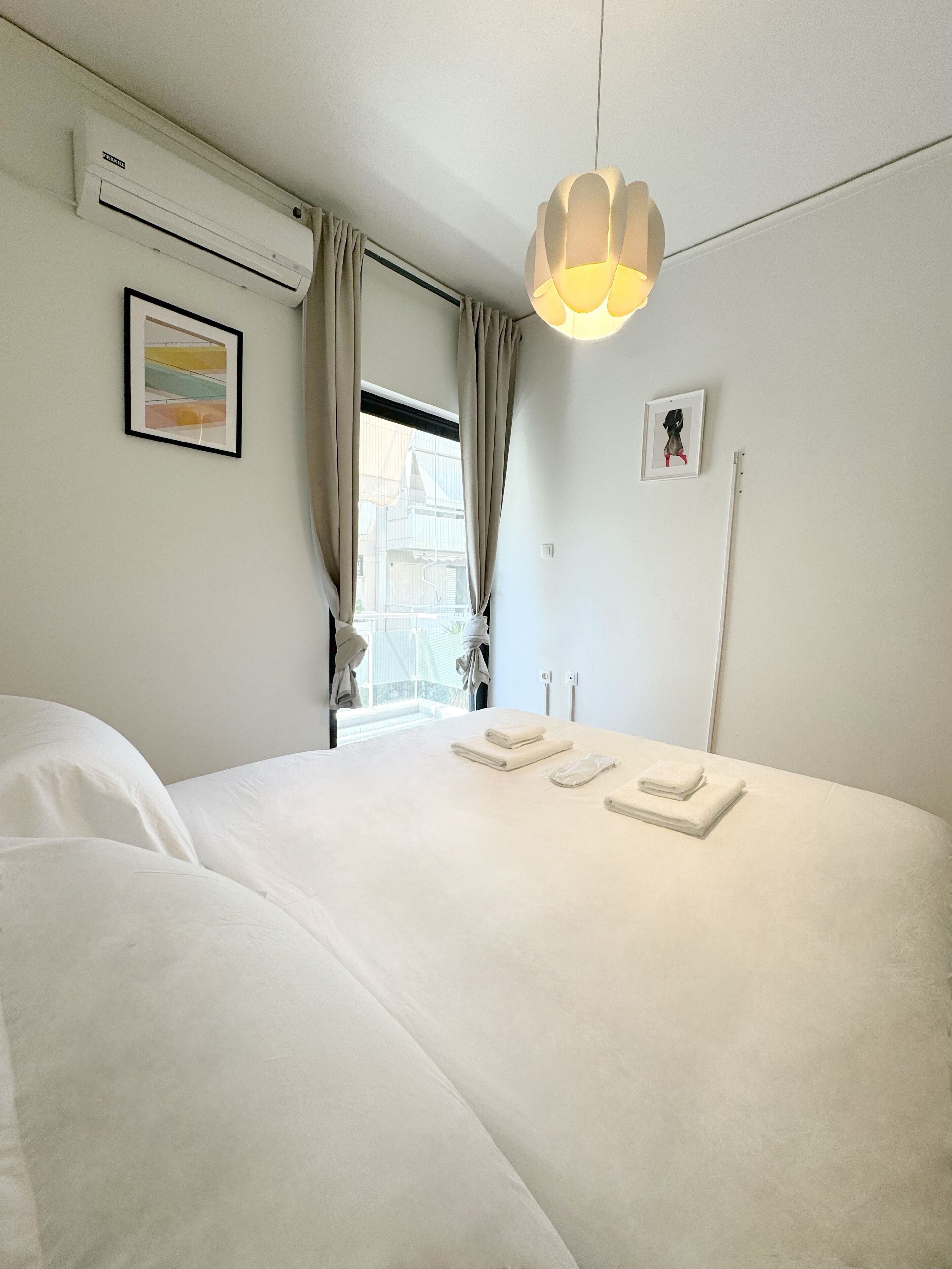 A hotel room with a large bed, an air conditioning unit, a hanging light fixture, and a window with curtains.