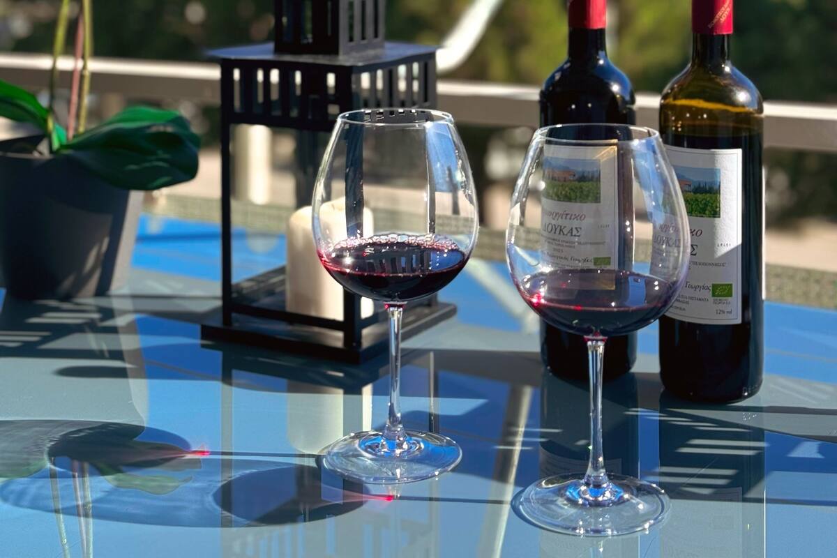 A glass table with two wine glasses, one filled with red wine, and two wine bottles. There is a potted plant and a small black lantern on the table.
