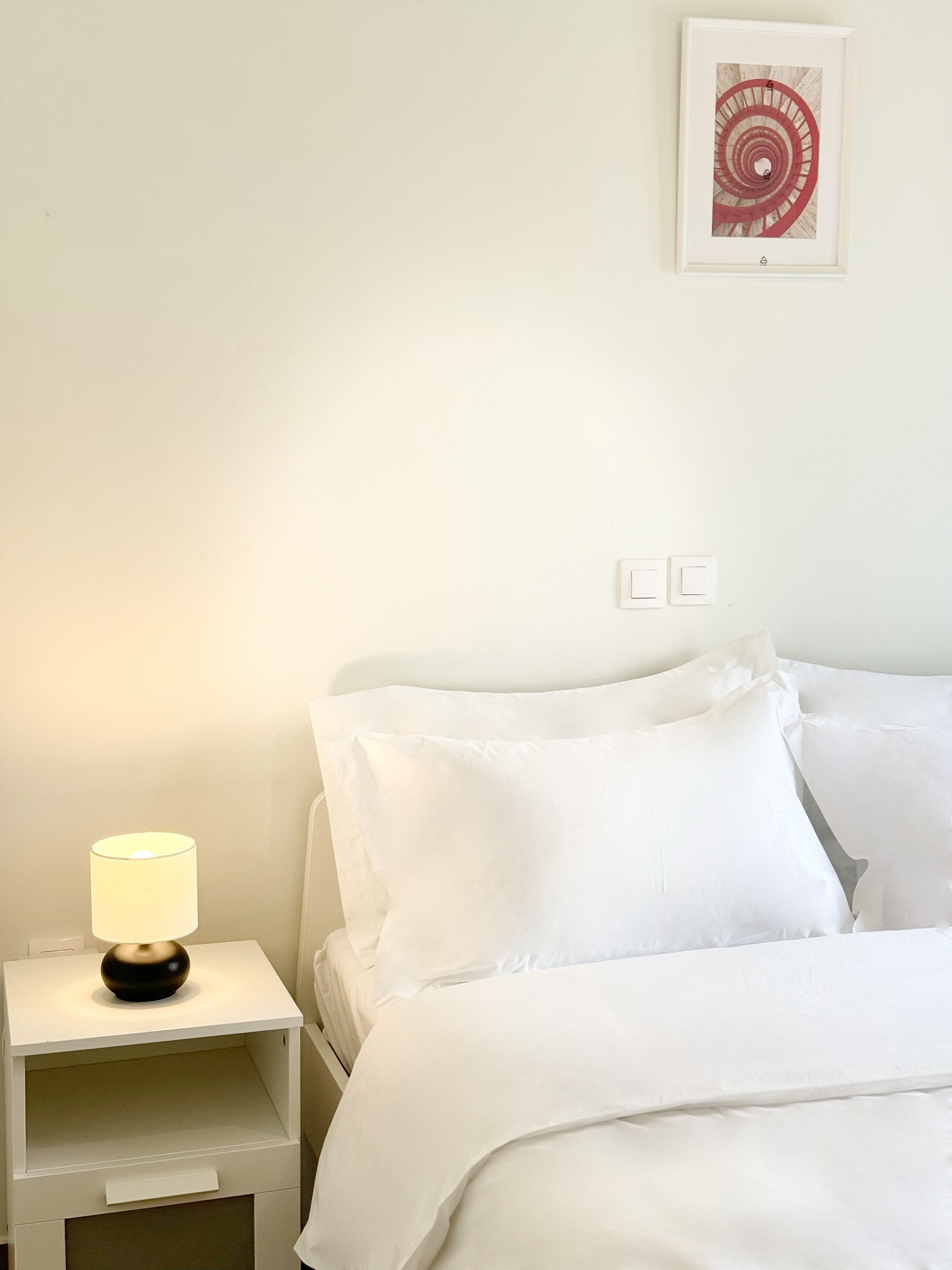 Ein ordentlich gemachtes Bett mit weißer Bettwäsche, ein Nachttisch mit Lampe und ein gerahmtes Bild an der Wand.
