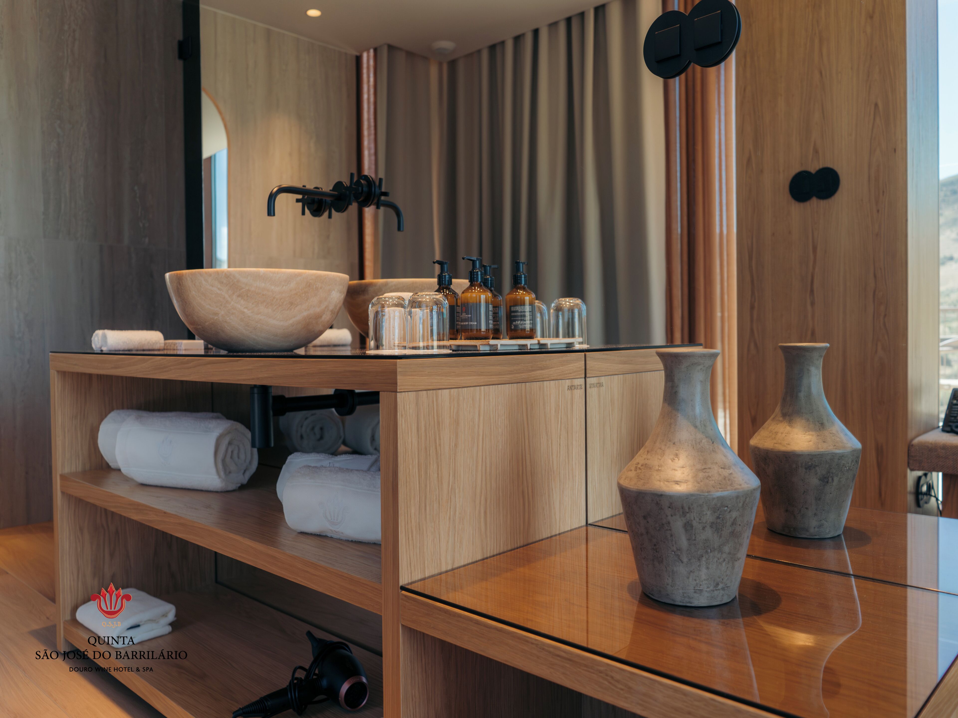 Un ba ño moderno con un mueble de tocador de madera, un lavabo grande y dos jarrones. Hay toallas y artículos de aseo en el mueble.