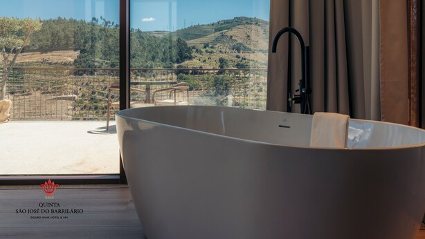 A modern bathroom with a freestanding bathtub, a large window offering a view of a vineyard, and a visible logo for Quinta São José do Barrilário.