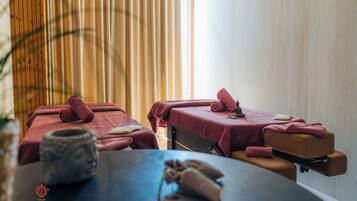 Sala de spa com duas mesas de massagem cobertas com toalhas vermelhas, uma bacia de pedra e uma parede de madeira.