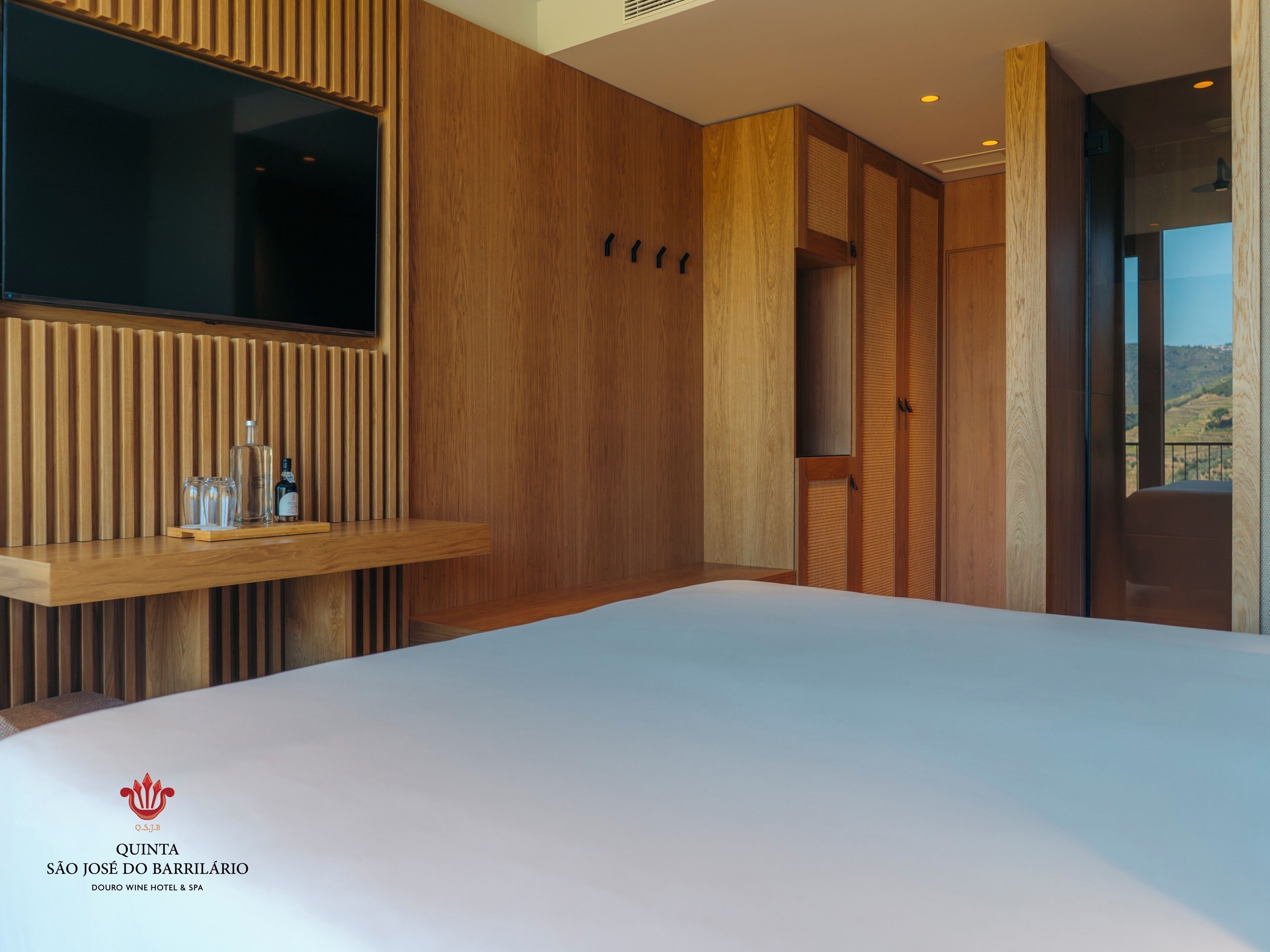 Una habitación de hotel moderna con un panel de pared de madera, un televisor montado, una estantería de madera con vasos y botellas, y una cama.