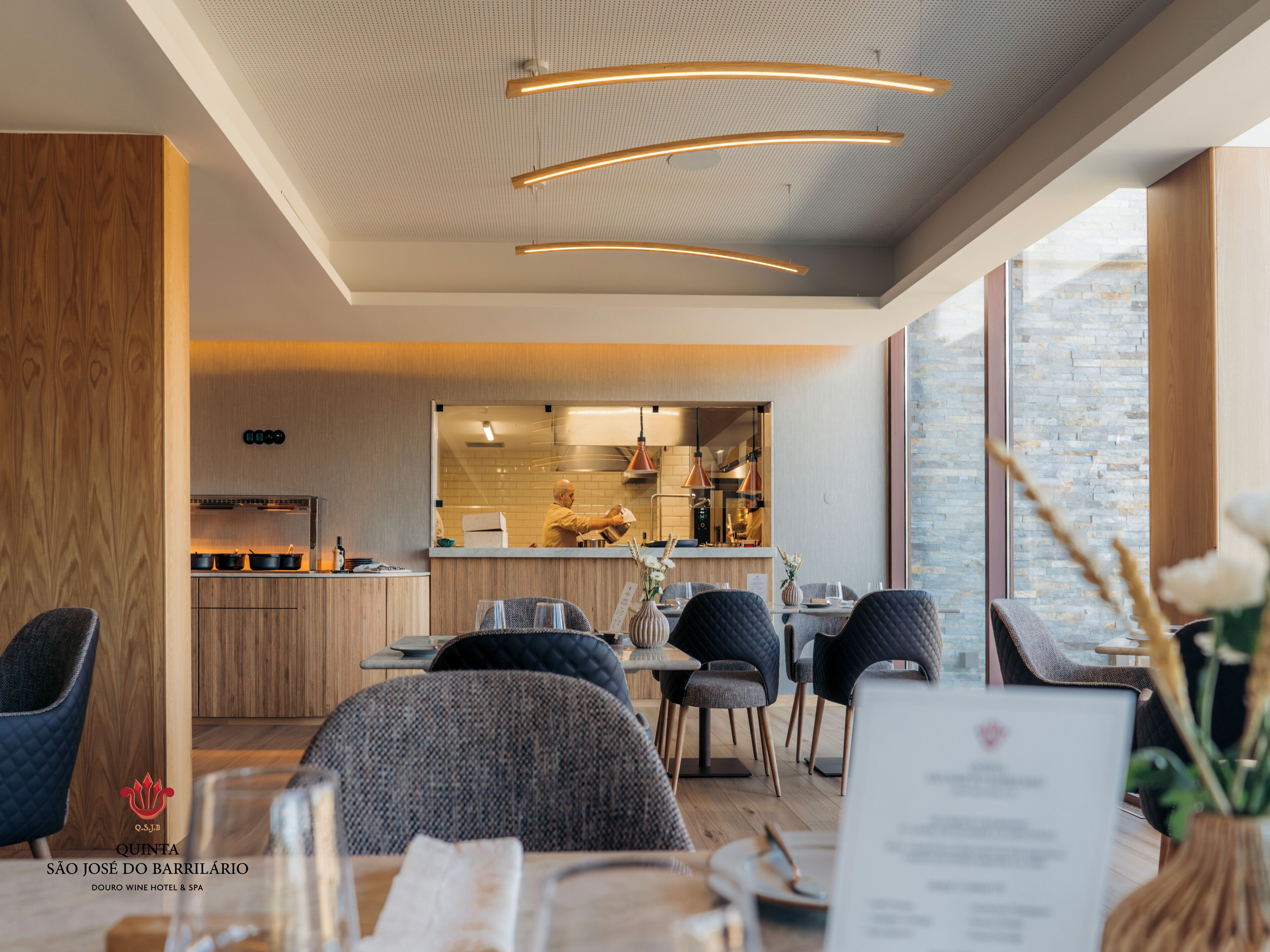 A modern dining area with a kitchen in the background, wooden accents, and large windows.