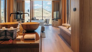 A modern hotel room with a large wooden dining table, two wooden bowls, a flat-screen TV, and a balcony with a scenic view.