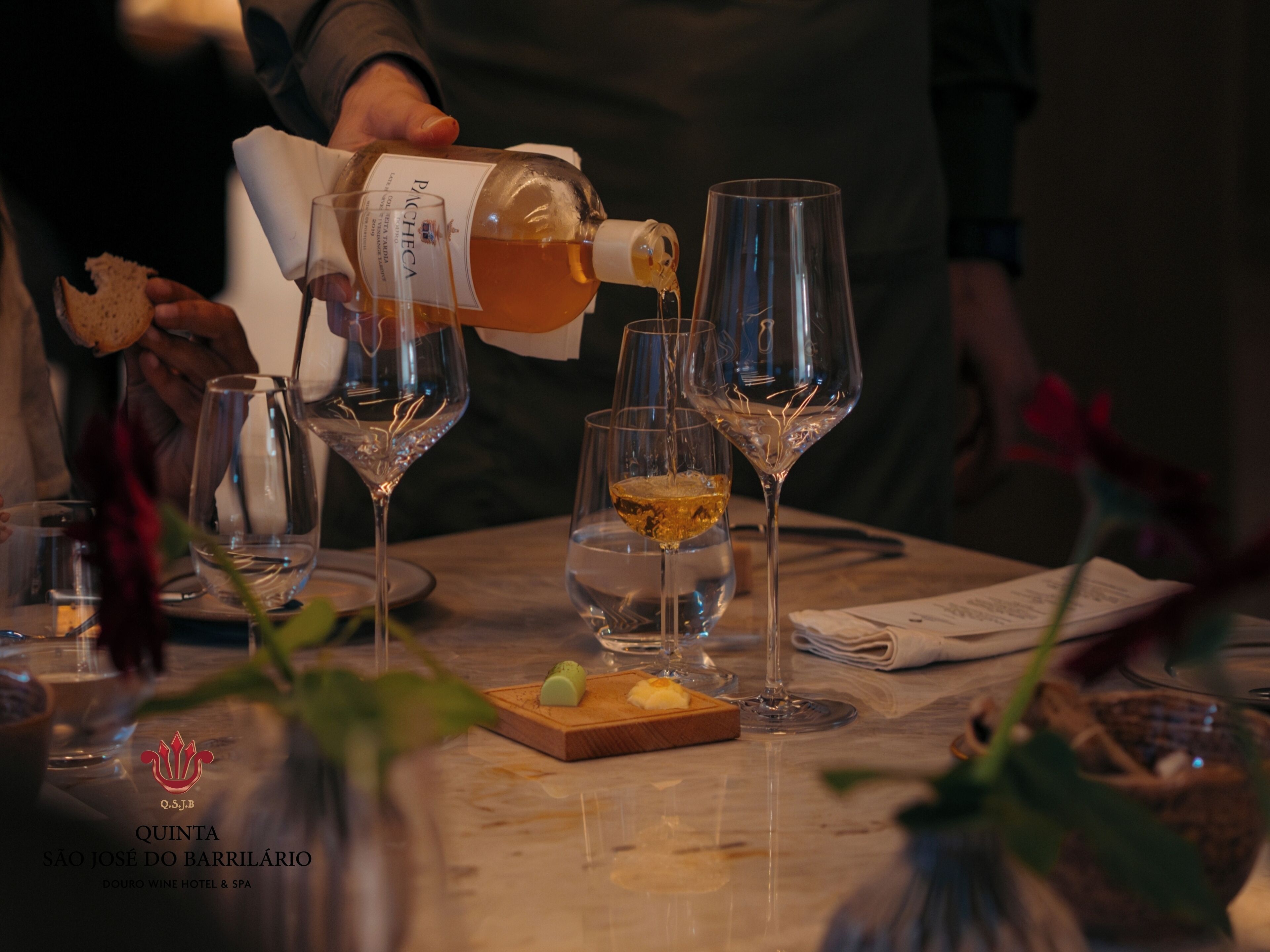 A wine tasting event with several wine glasses on a table, a bottle of wine being poured, and a wooden board with a small dish.
