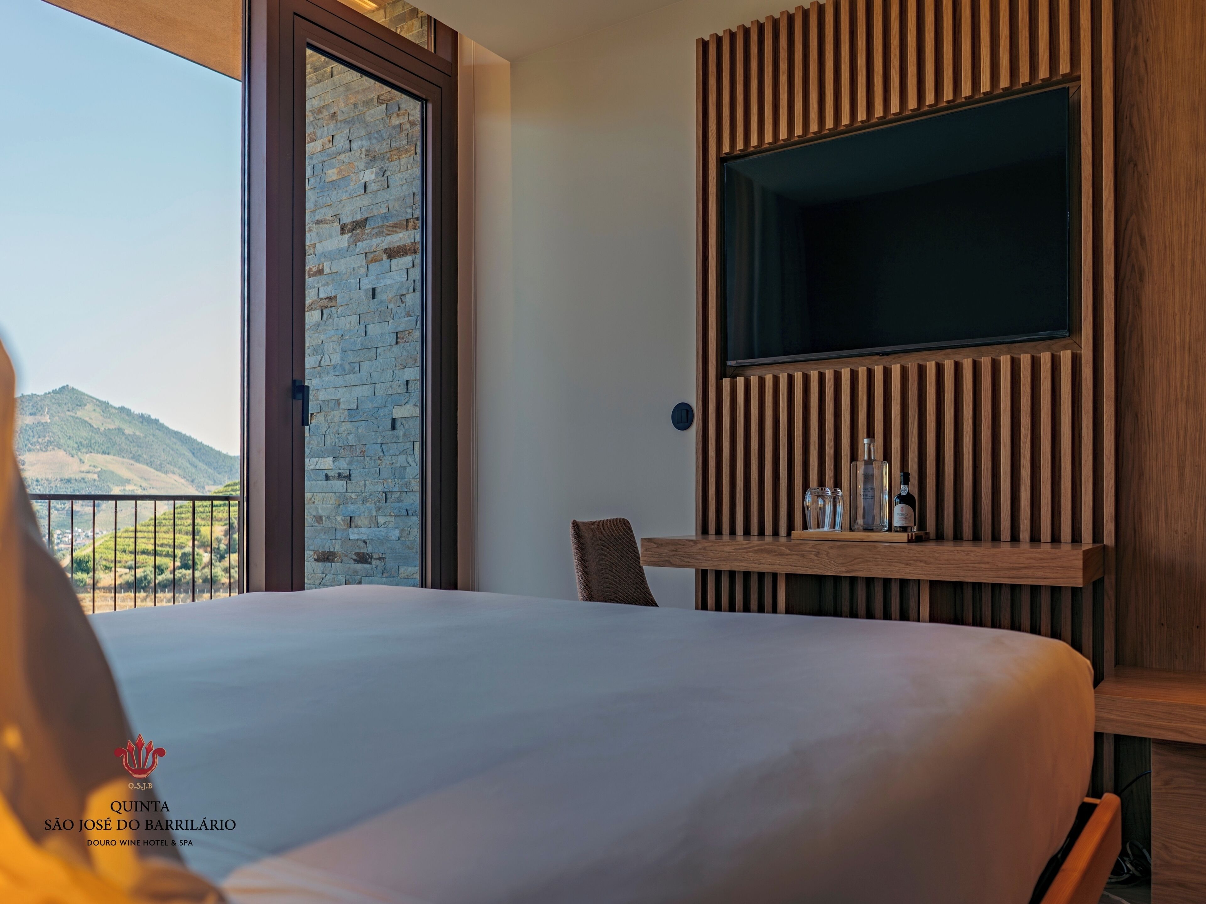 Habitación de hotel con una cama grande, un televisor montado en un panel de madera, una mesa de vidrio con botellas y vistas a la montaña a través de una puerta de cristal.