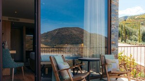 A balcony with two wicker chairs, a small round table, and a view of a mountainous landscape through large glass doors.