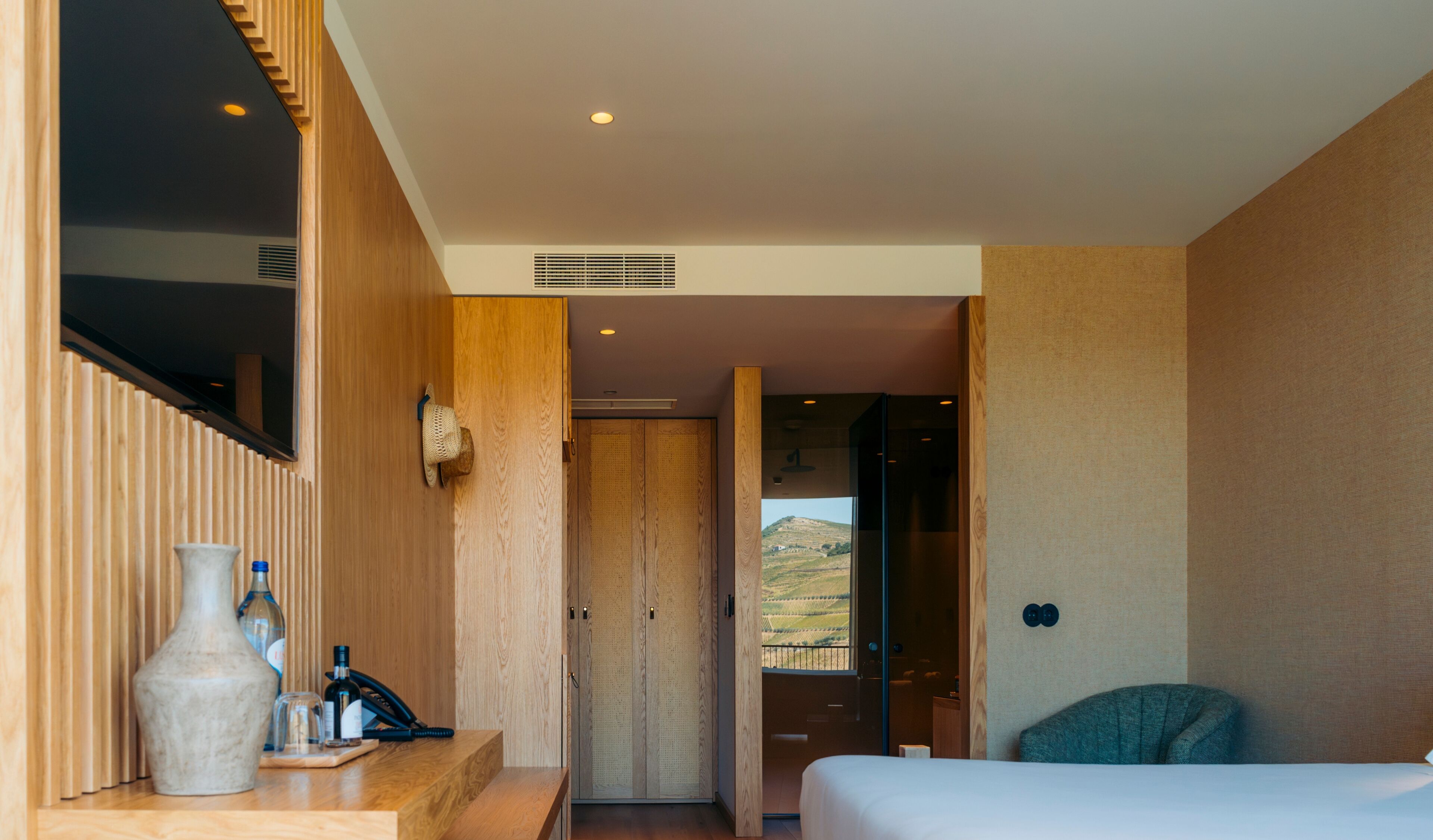 Habitación de hotel moderna con una pared decorada en madera, un ventanal con vistas a un paisaje montañoso, una cama y un escritorio con un florero y botellas.
