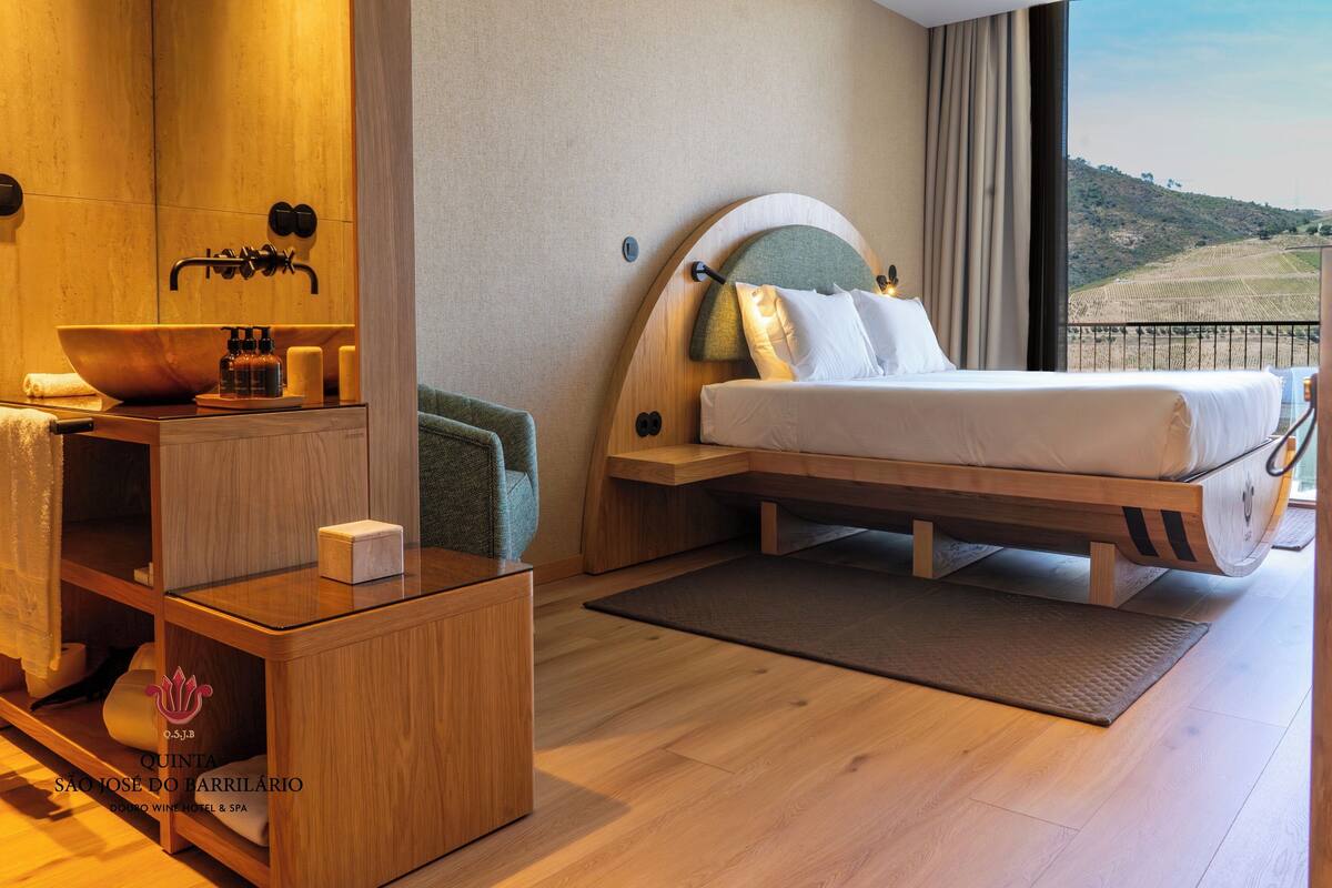 Ett modernt hotellrum med en stor säng, ett träsminkbord med handfat och utsikt över en vingård genom ett stort fönster.