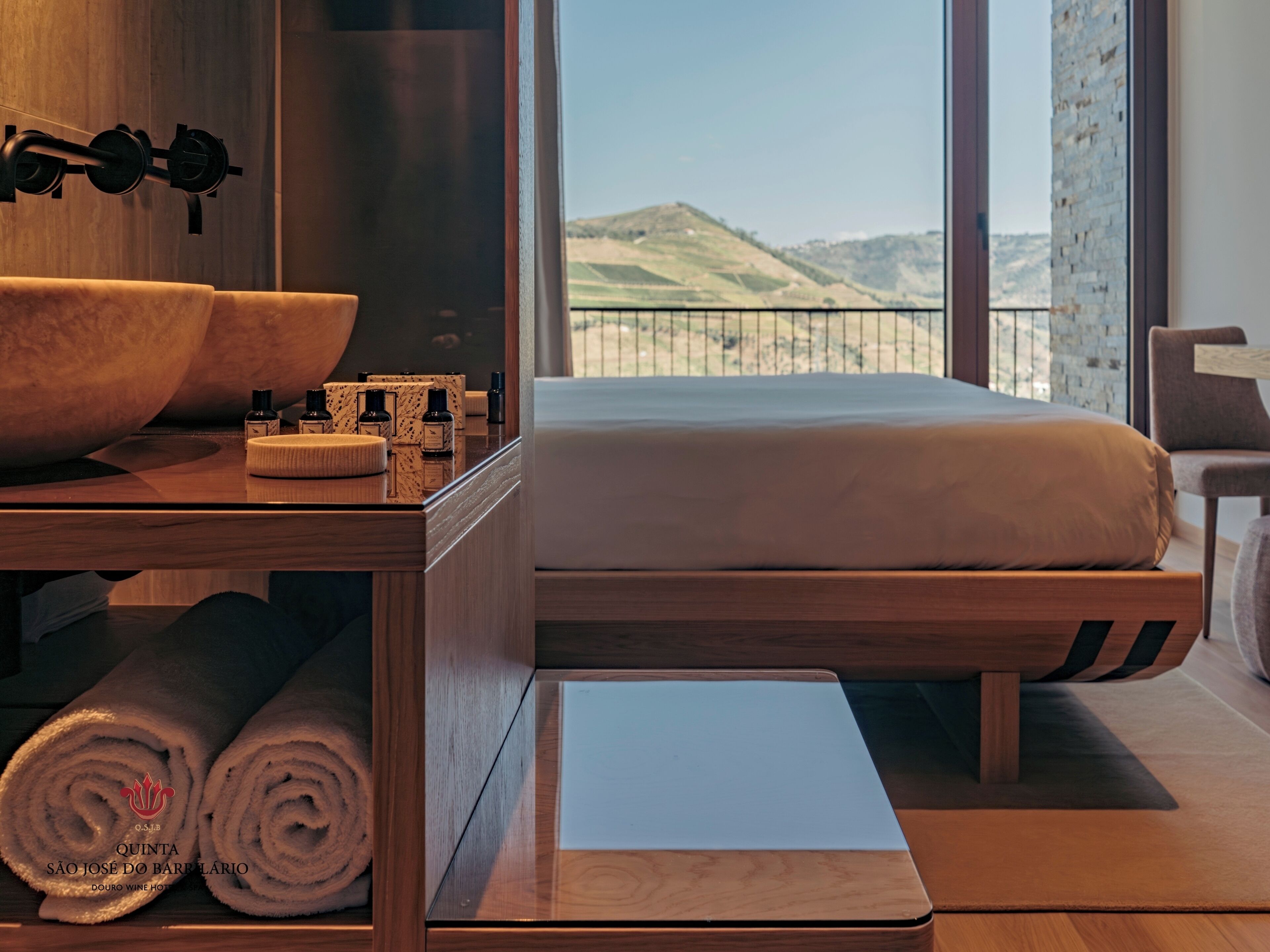Quarto de hotel moderno com cama de madeira, uma janela ampla com vista para colinas, uma mesa de cabeceira de madeira com artigos de higiene e uma pia grande.