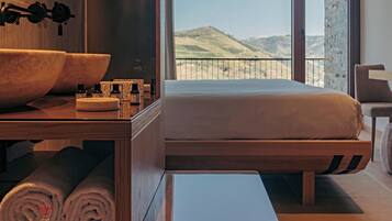 Quarto de hotel moderno com cama de madeira, uma janela ampla com vista para colinas, uma mesa de cabeceira de madeira com artigos de higiene e uma pia grande.