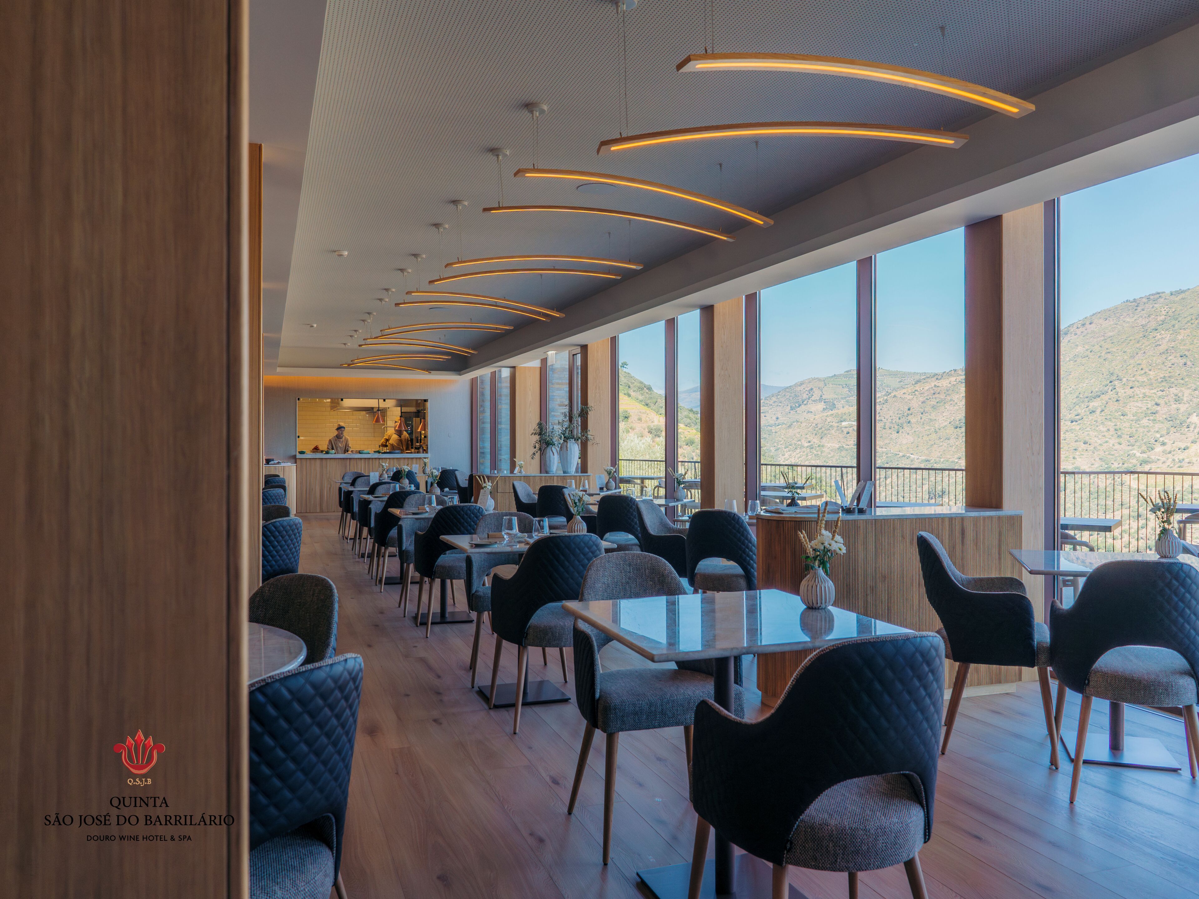 A modern dining area with tables and chairs, large windows offering a view of the hills, and a visible kitchen area.