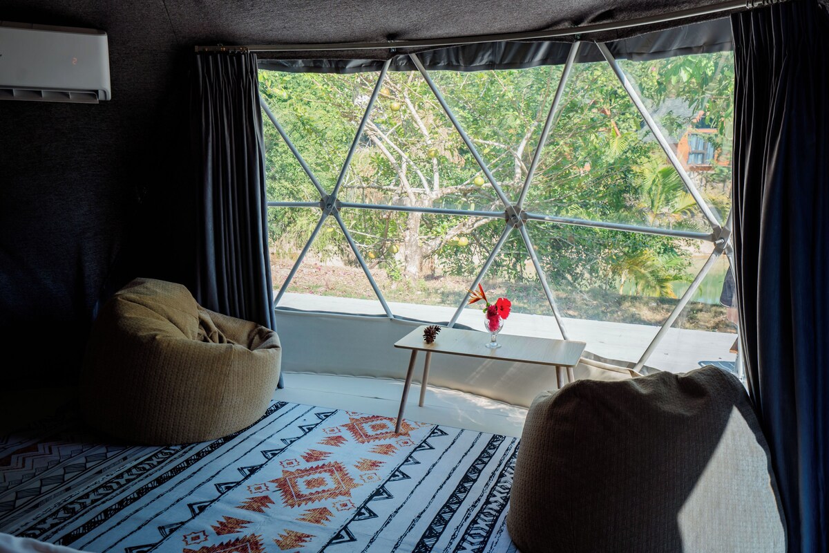 A room with a large window, a bed, a small table with a vase, and a bean bag chair.
