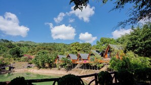 A row of wooden chalets with sloped roofs, built on stilts over a body of water, surrounded by lush greenery and hills.