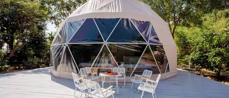 A geodesic dome structure with a transparent covering, surrounded by a deck with white chairs and a table, set in a lush, green environment.
