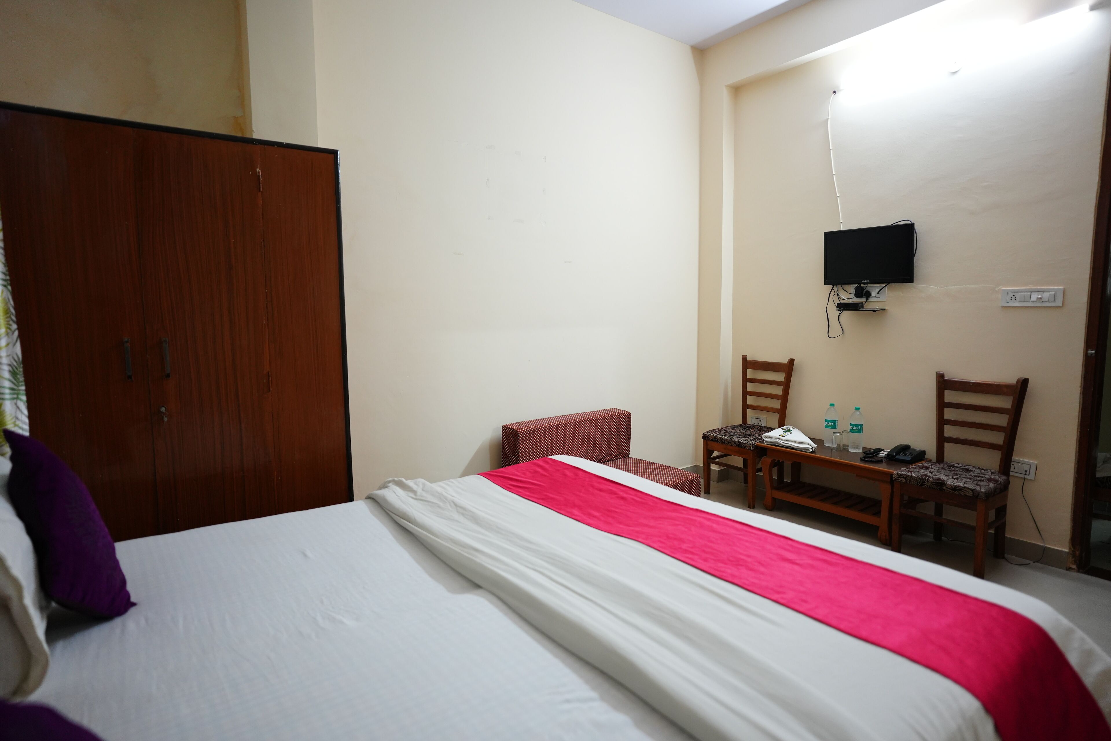 A hotel room with a bed, a wooden wardrobe, a television mounted on the wall, and a small table with chairs.