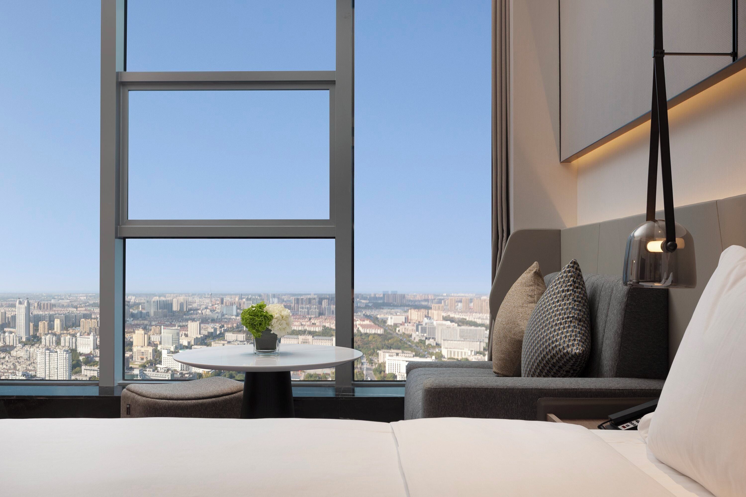 A modern hotel room with a large window offering a city view, a round table with a plant, a sofa with cushions, and a hanging lamp.