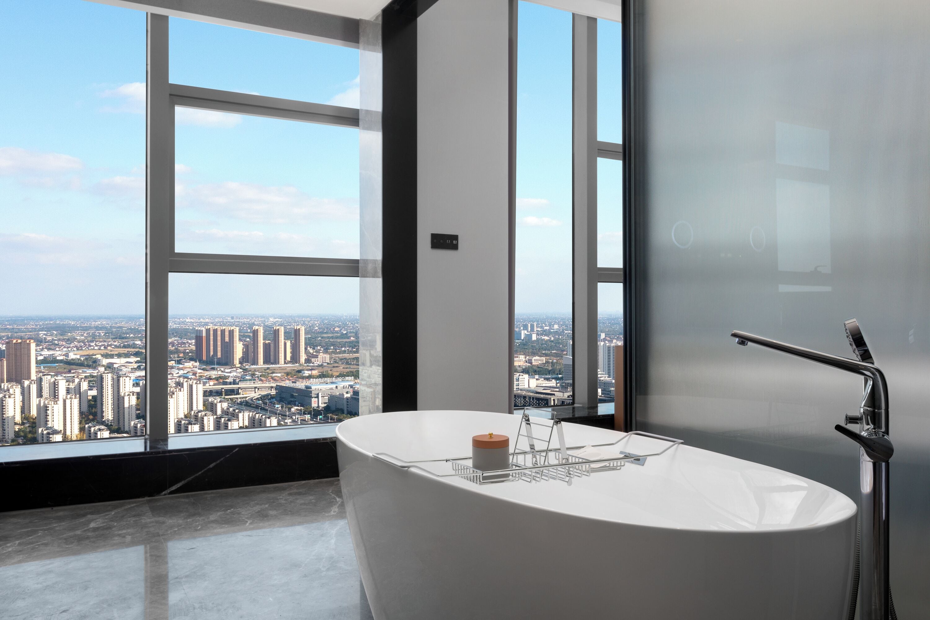 A modern bathroom with a freestanding bathtub, a large window offering a city view, and a frosted glass door.