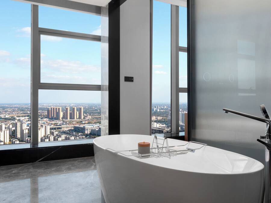A modern bathroom with a freestanding bathtub, a large window offering a city view, and a frosted glass door.