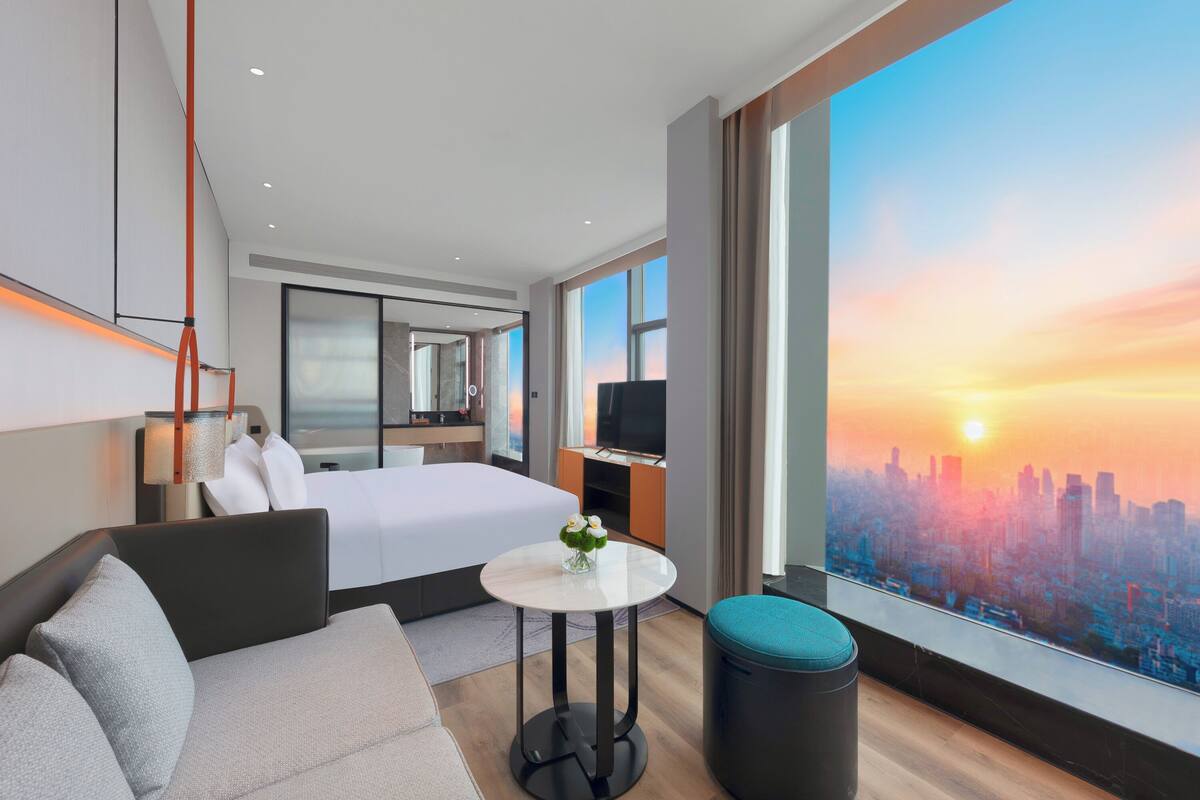 A modern hotel room with a bed, sofa, round table, and a large window with a city view.