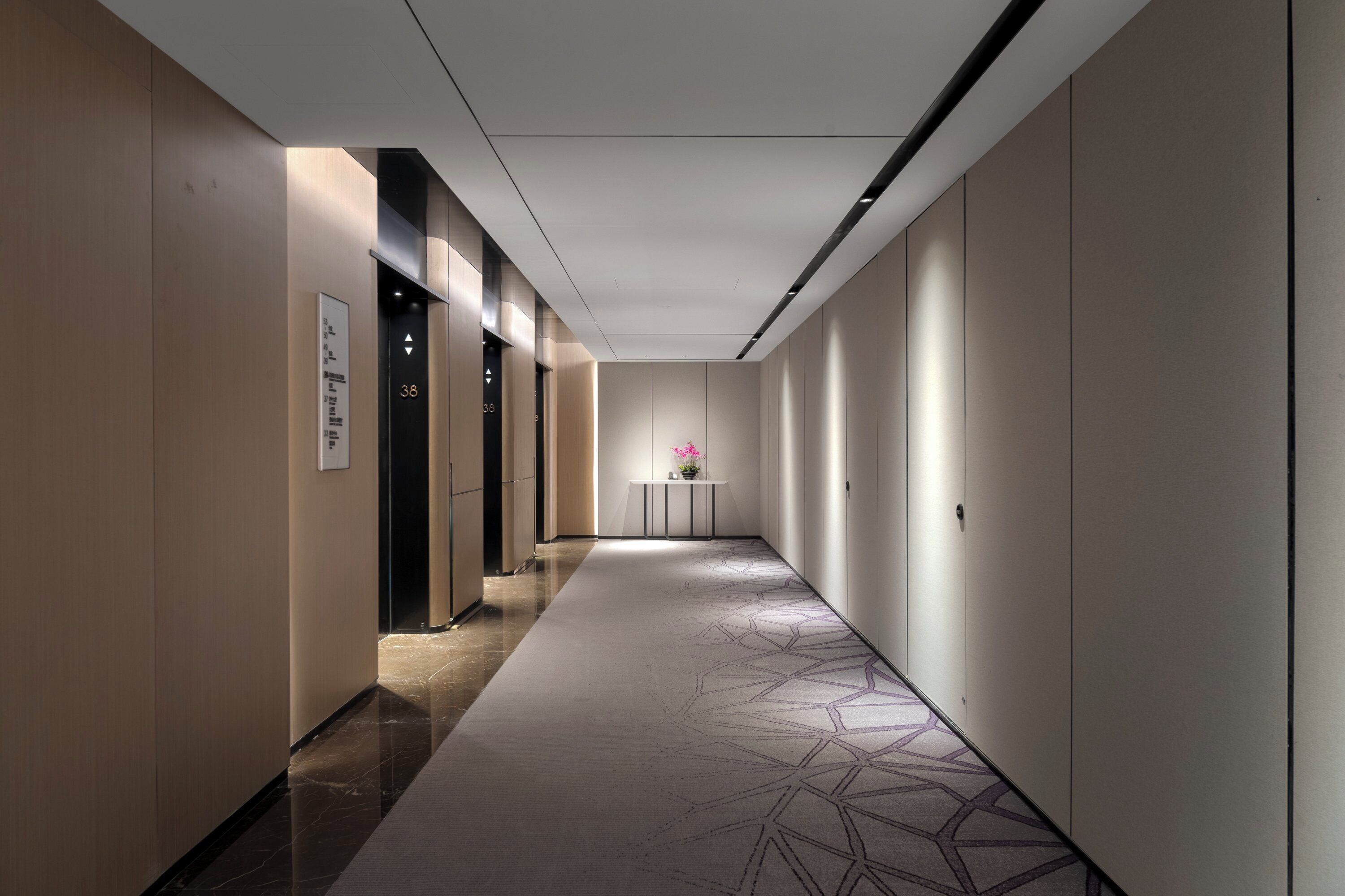 Un pasillo moderno de hotel con una alfombra de diseño geométrico, un mostrador de recepción y una señal en la pared que indica la habitación número 38.