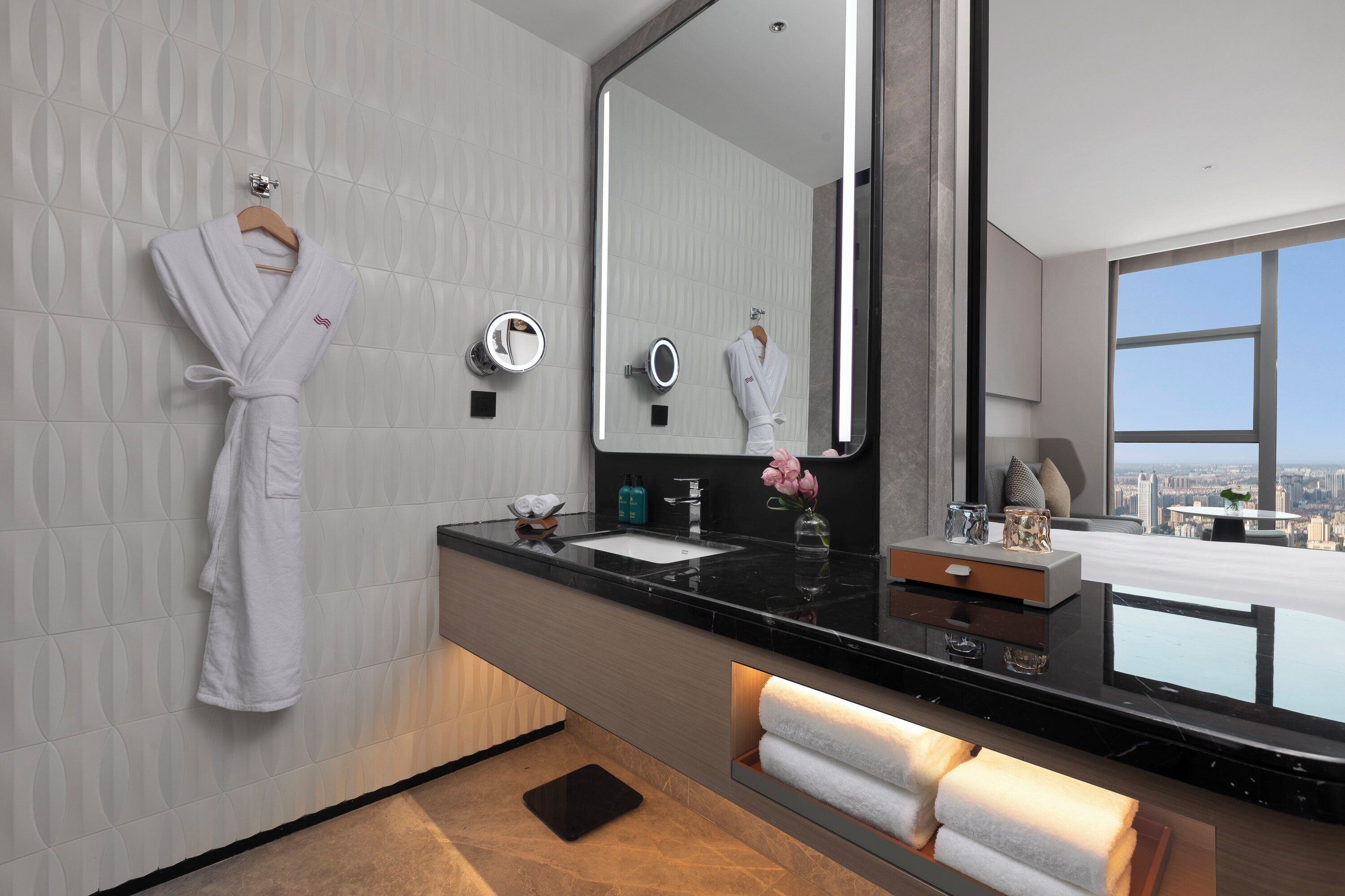 Een moderne badkamer met een grote spiegel, twee wastafels, een badjas hangend aan de muur en uitzicht op de stad door het raam.