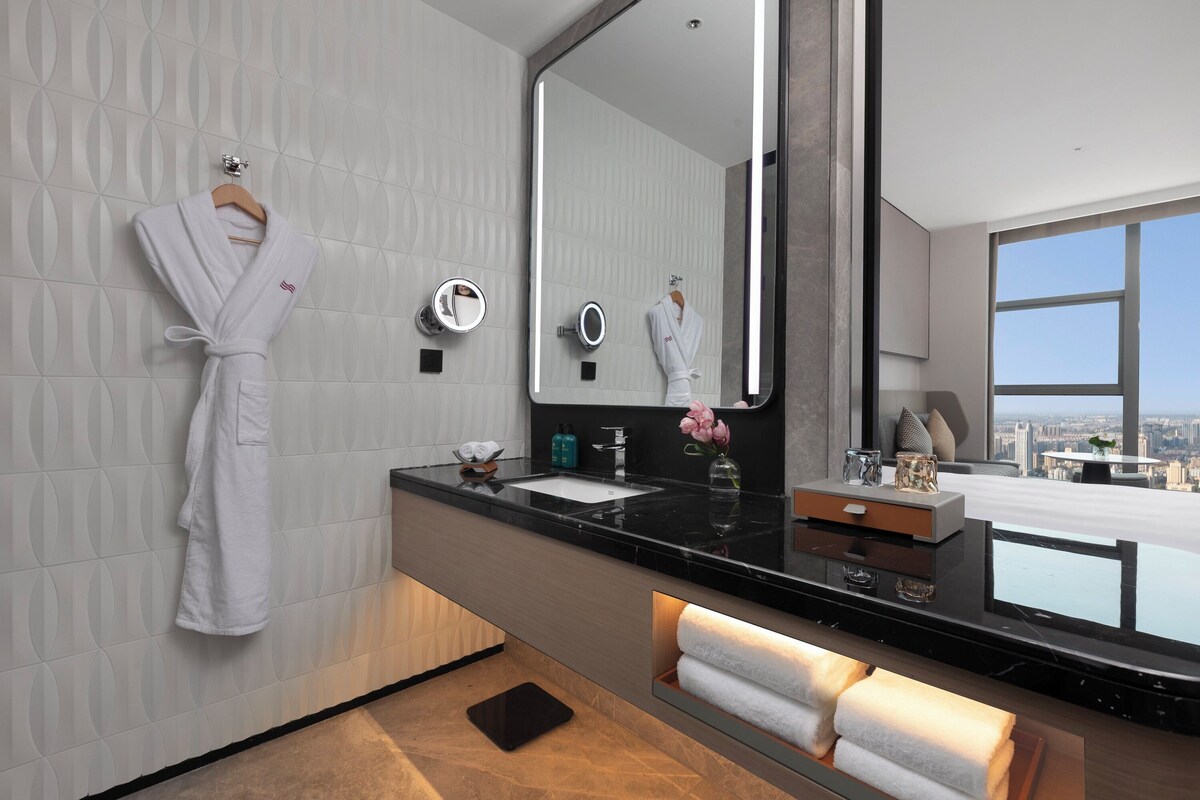 A modern bathroom with a large mirror, two sinks, a robe hanging on the wall, and a view of the city through the window.