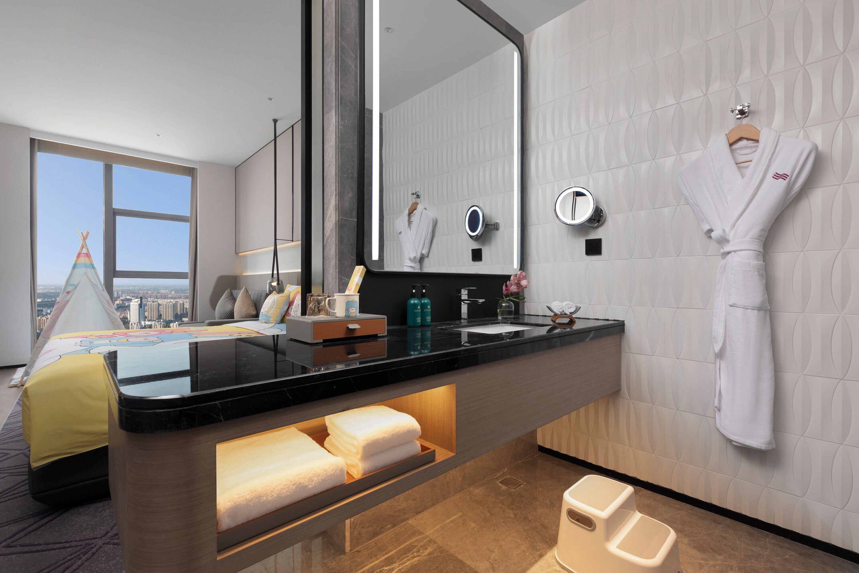 A modern hotel bathroom with a large mirror, a vanity with a black countertop, and a white robe hanging on the wall.