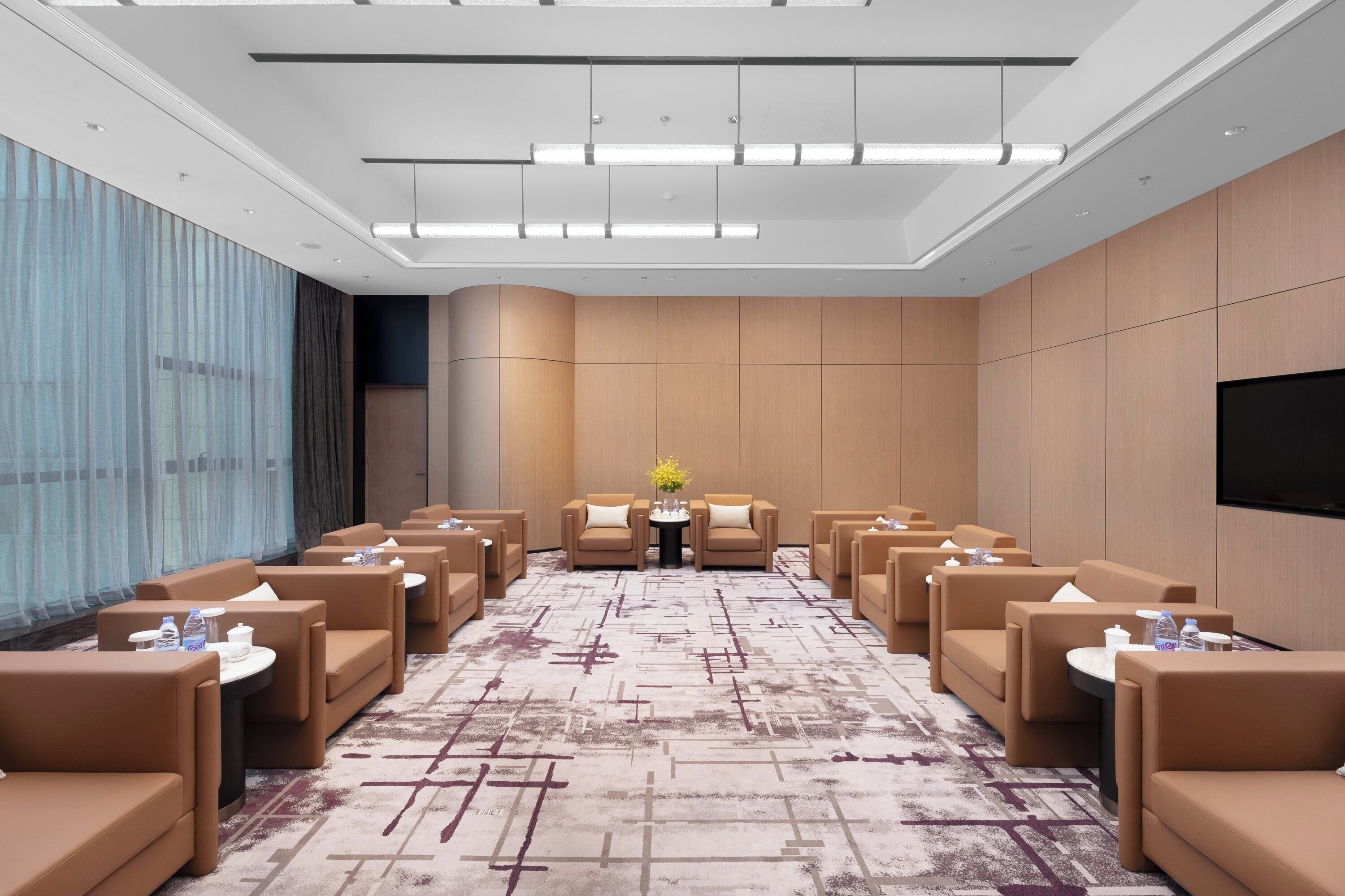A hotel conference room with rows of brown sofas, a central table with a vase of flowers, and a flat-screen TV on the wall.
