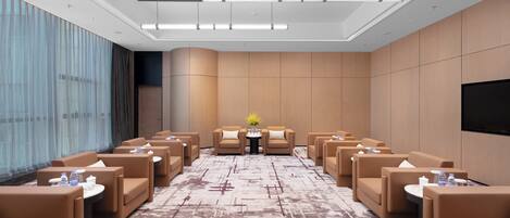A hotel conference room with rows of brown sofas, a central table with a vase of flowers, and a flat-screen TV on the wall.