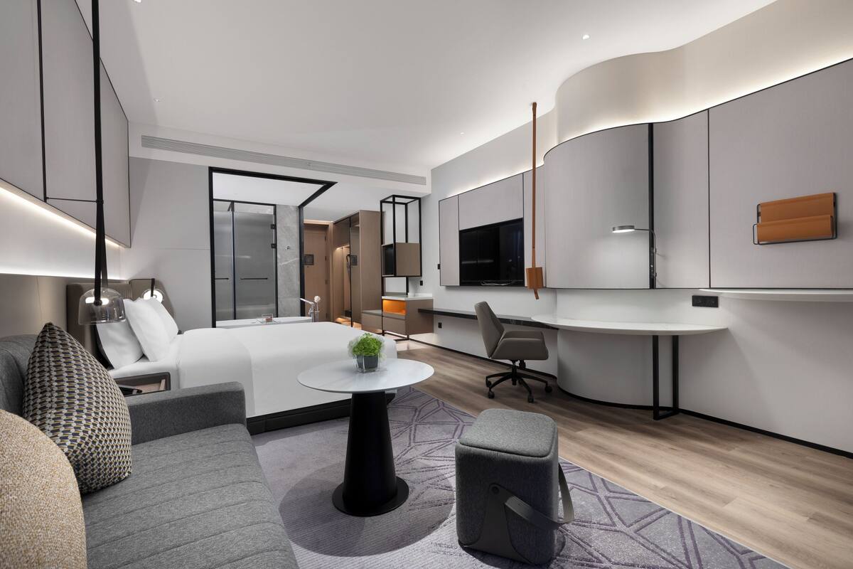 A modern hotel room with a bed, a desk, a sofa, and a small table with a plant.