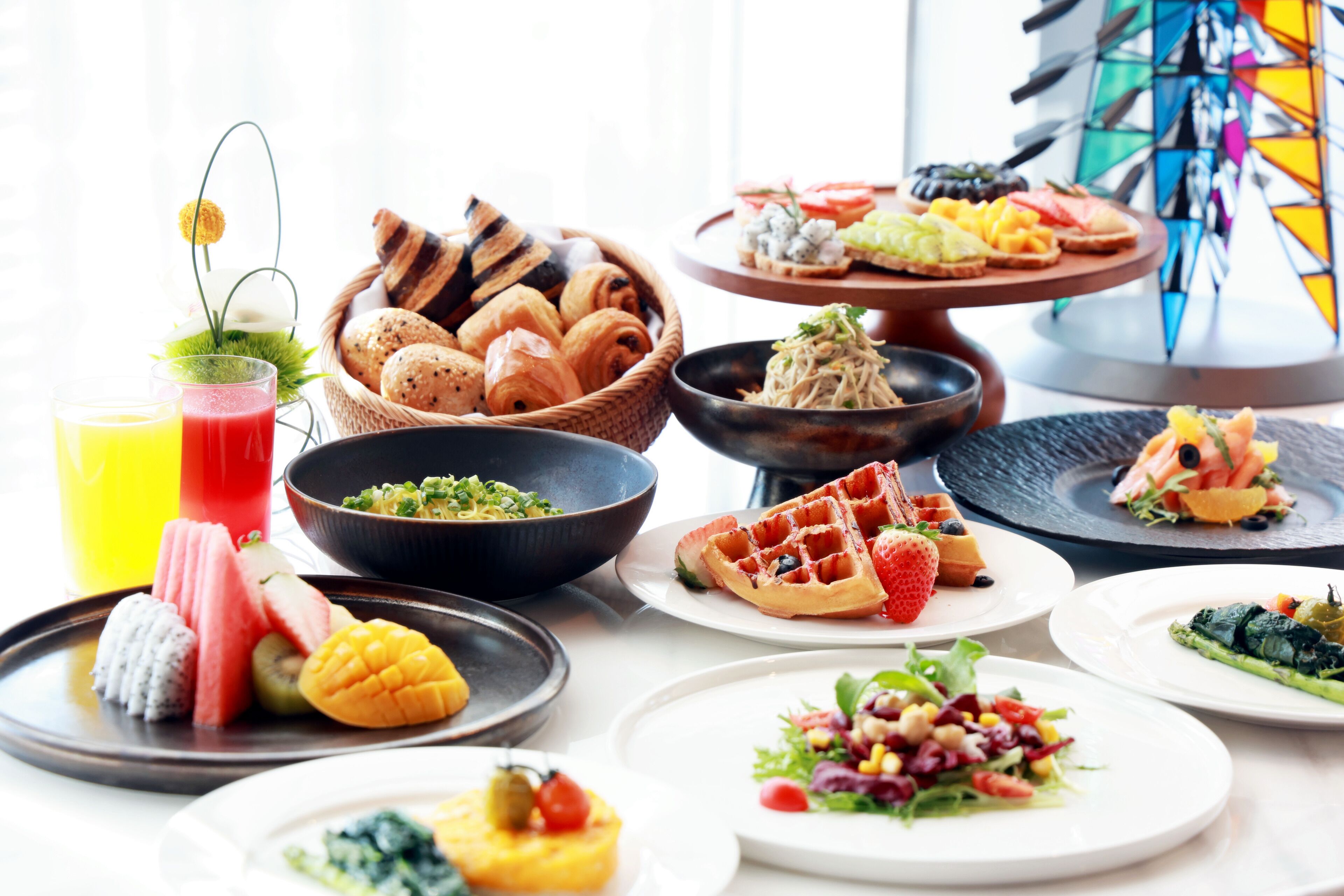 Un assortiment de produits pour le petit-déjeuner, notamment des viennoiseries, des gaufres et des fruits présentés sur des assiettes, avec en arrière-plan un vitrail aux couleurs vives.